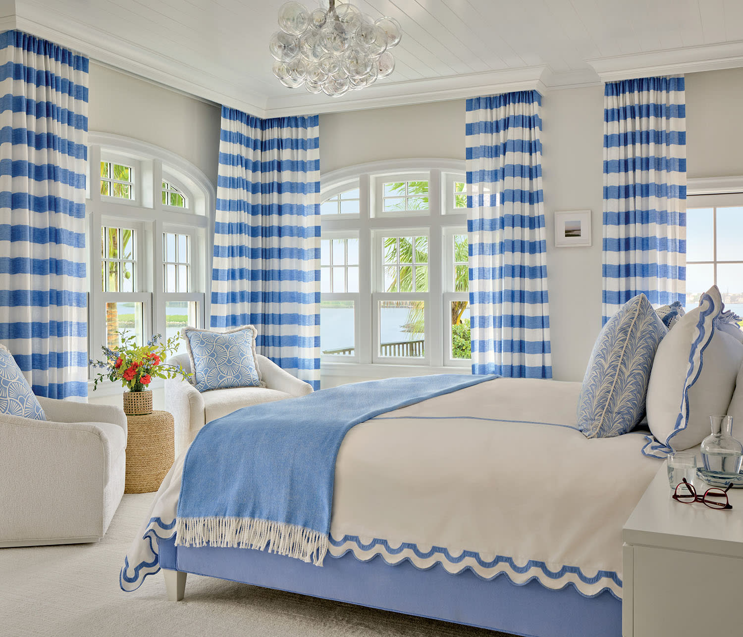 A serene bedroom with coastal decor, featuring blue and white striped curtains, a cozy bed with light blue accents, and a soft armchair beside a flower vase.