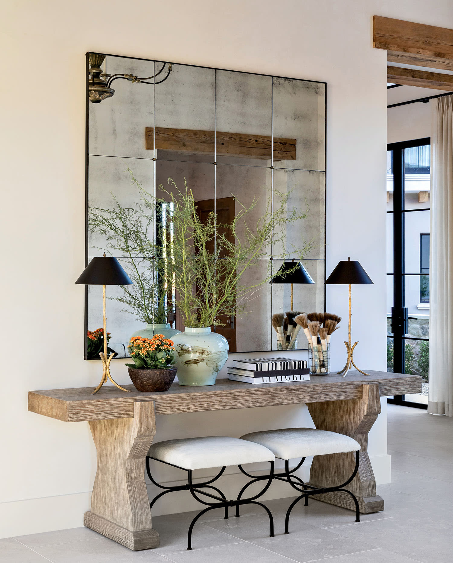Sophisticated interior with a rustic wood console, large mirror, and elegant decor like black lamps, a potted plant, and stacked books. Airy and calm ambiance.