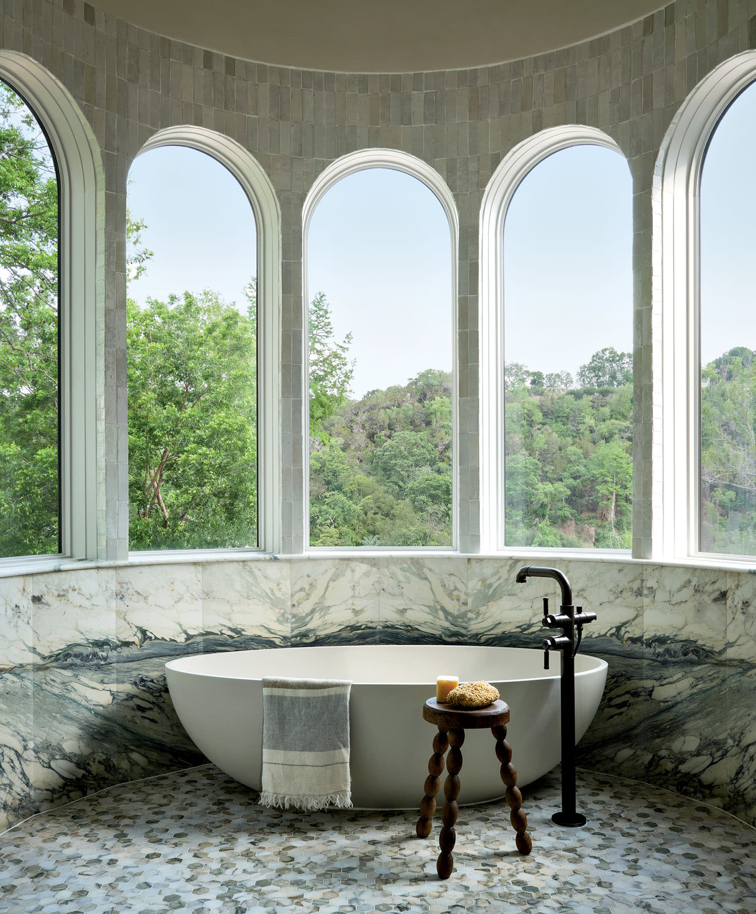 Luxurious bathroom with a white freestanding tub set against large arched windows, offering a scenic view of green trees. A small wooden stool with a candle and sponge adds a cozy touch.