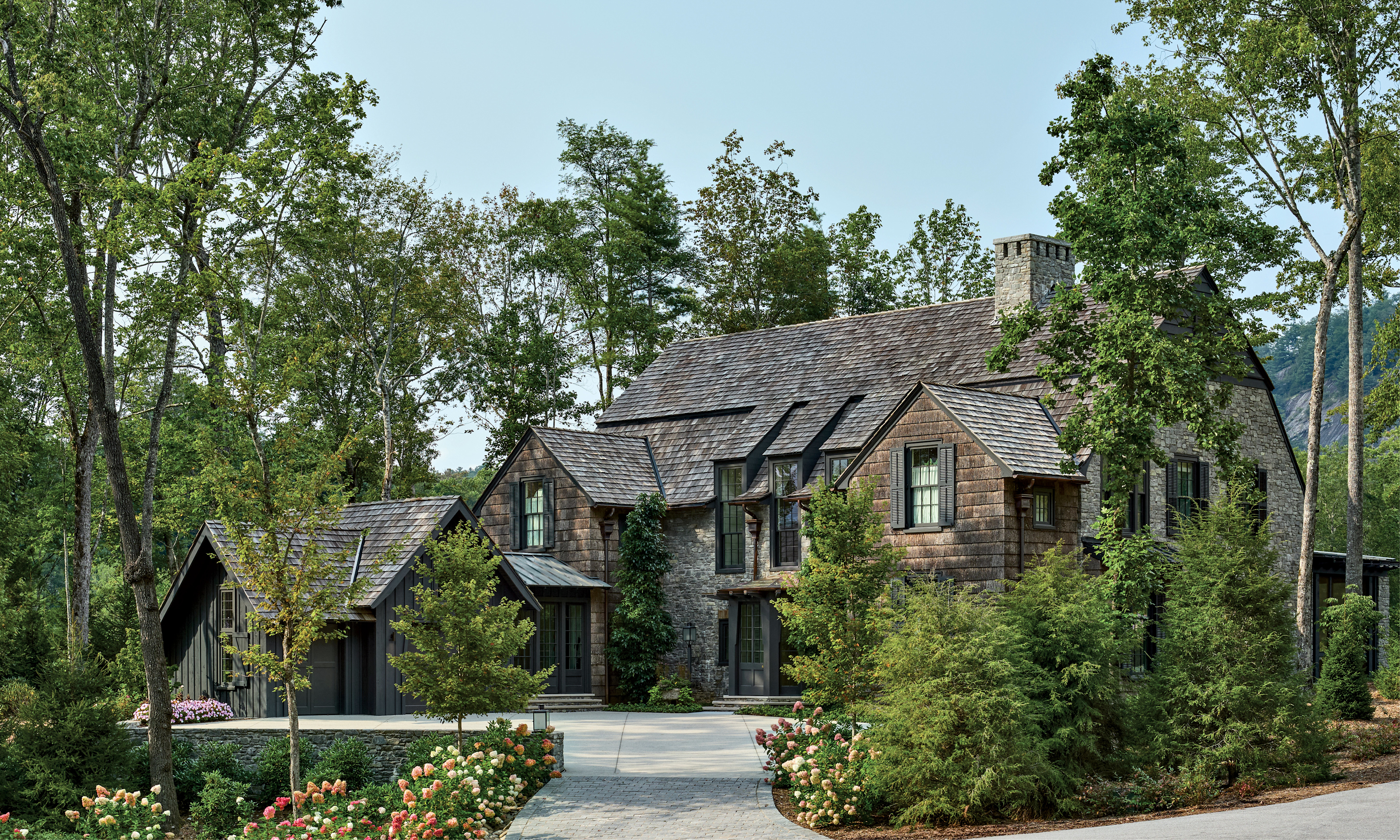 Stone country house surrounded by lush trees and colorful gardens under a clear sky. A curved driveway leads to the entrance, evoking tranquility.