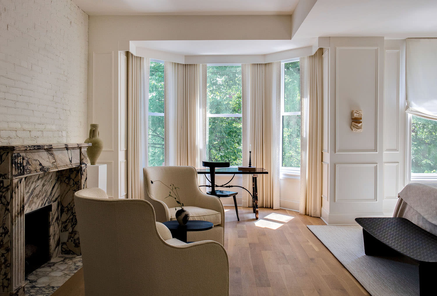 A bench sits at the edge of a bed opposite two chairs, near a desk and bay window looking out at trees.