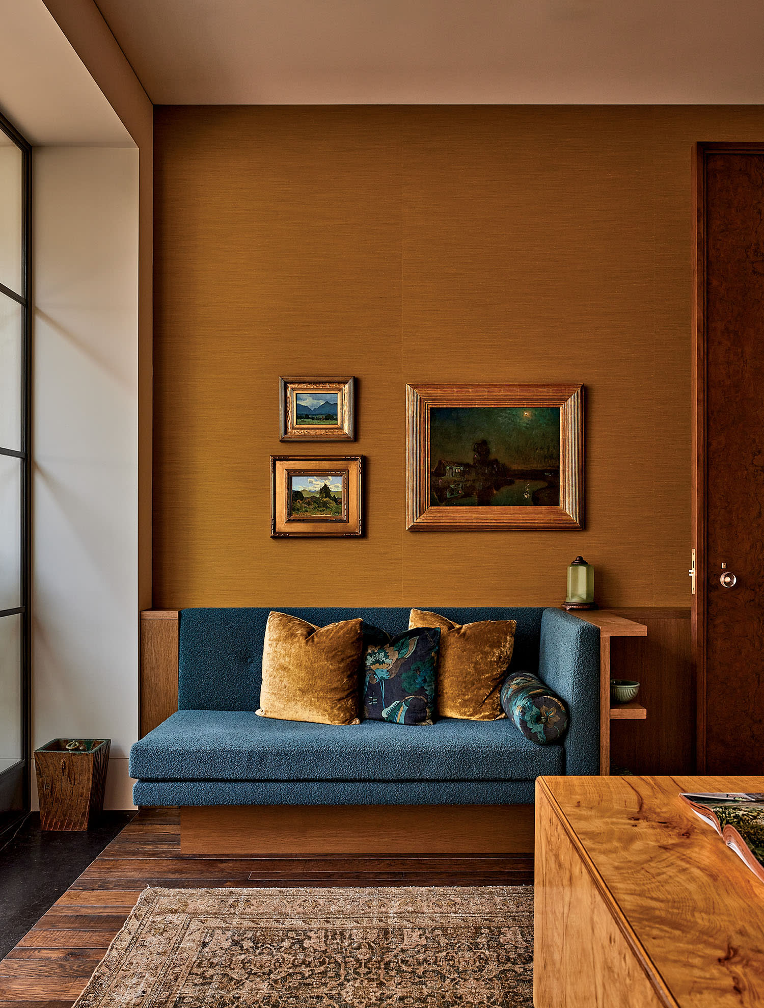 Cozy home office with a teal sofa, mustard cushions, and a warm brown wall. Framed landscape paintings add a classic touch above the couch.