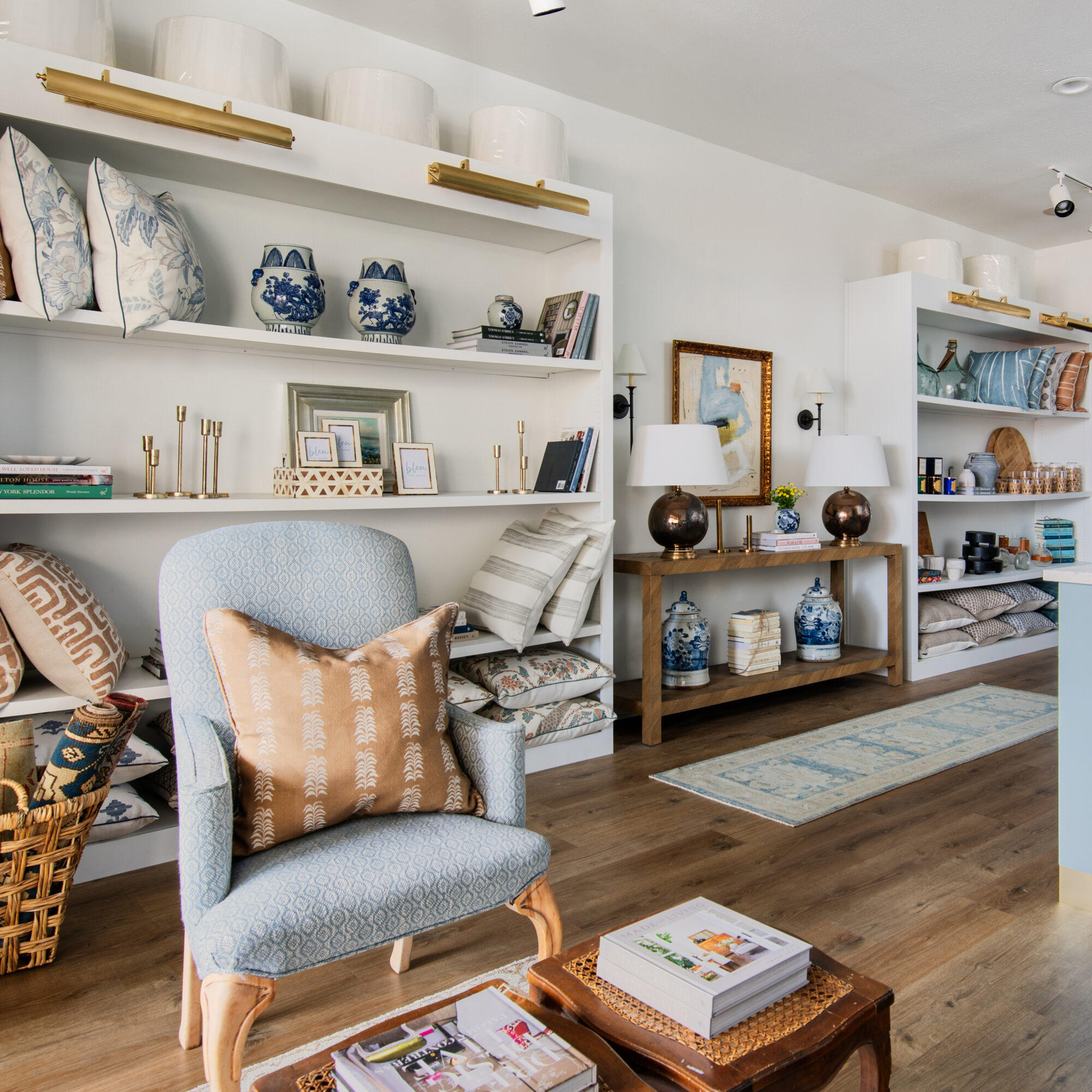 Wooden console table with lamps sits between large white shelving with pillows and home decor; a blue armchair sits atop wood floors.