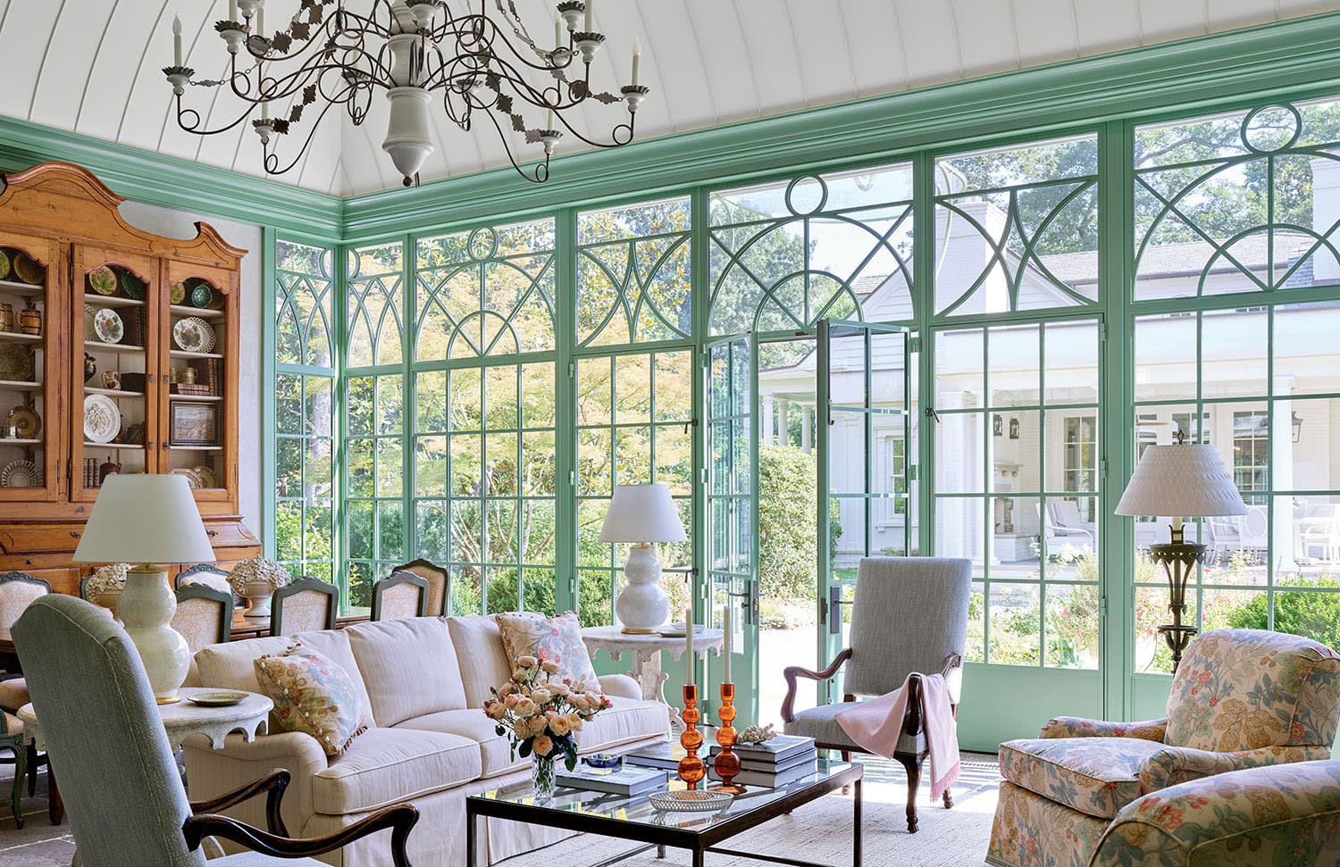 Elegant sunroom with large, green-framed windows, plush sofas, floral armchairs, and a vintage chandelier. Light-filled, cozy, and inviting atmosphere.