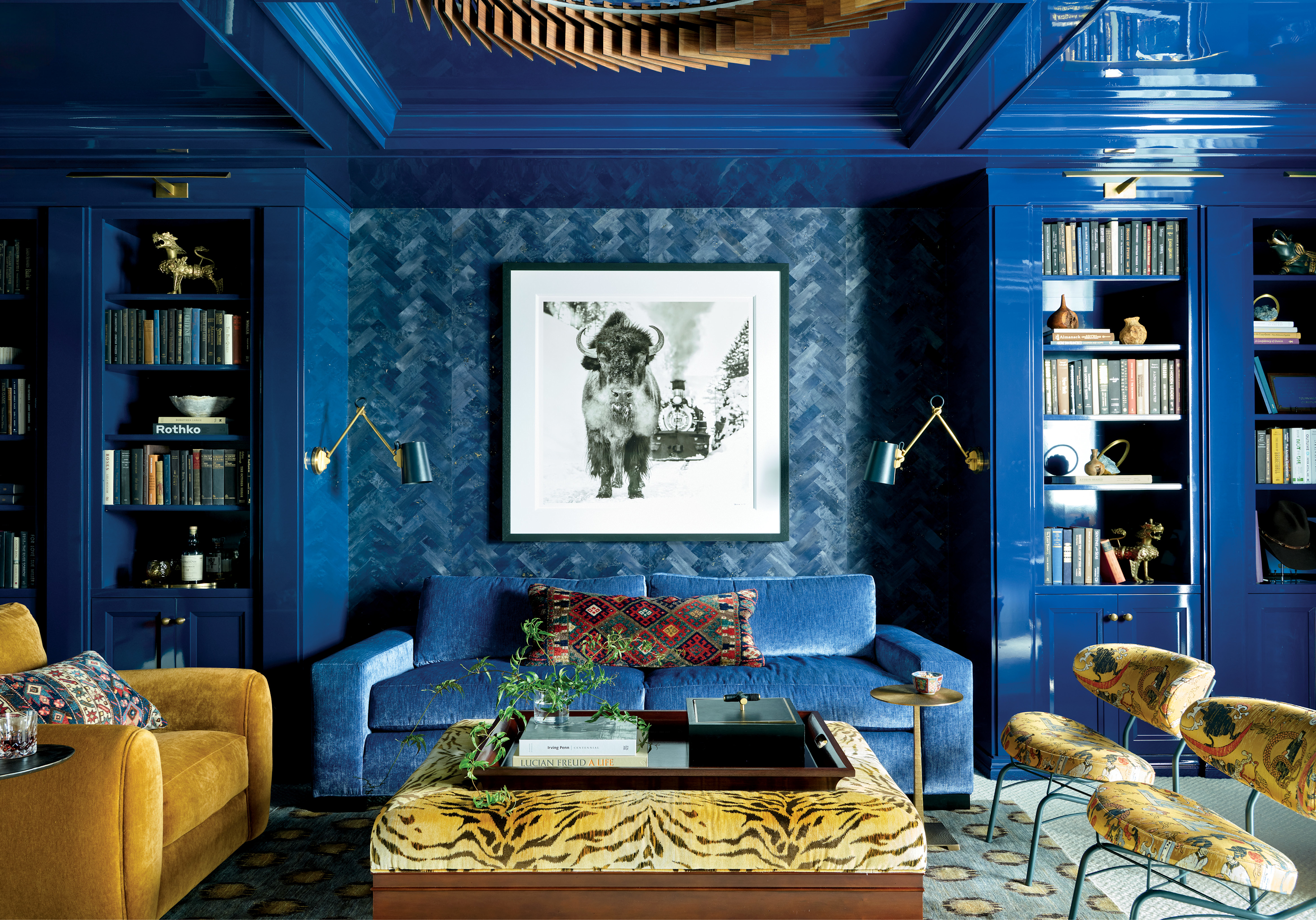 Vibrant living room with rich blue walls, bookshelves, and ceiling. Features a blue sofa, golden armchair, tiger-patterned ottoman, and a bison artwork.