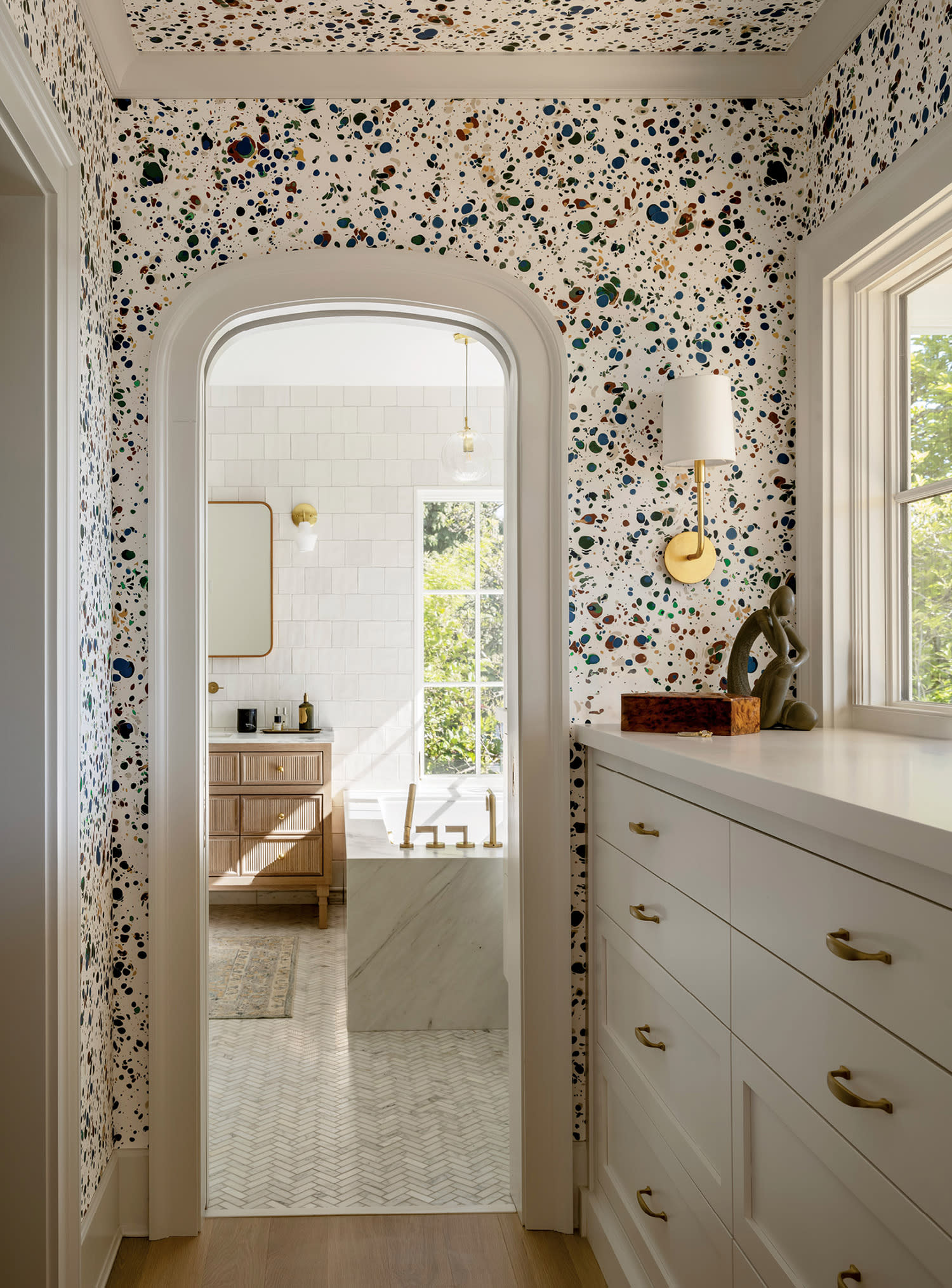 Narrow, brightly lit hallway with terrazzo-style speckled walls in white and colors. Leads to a sunlit bathroom with wood cabinets and large windows.