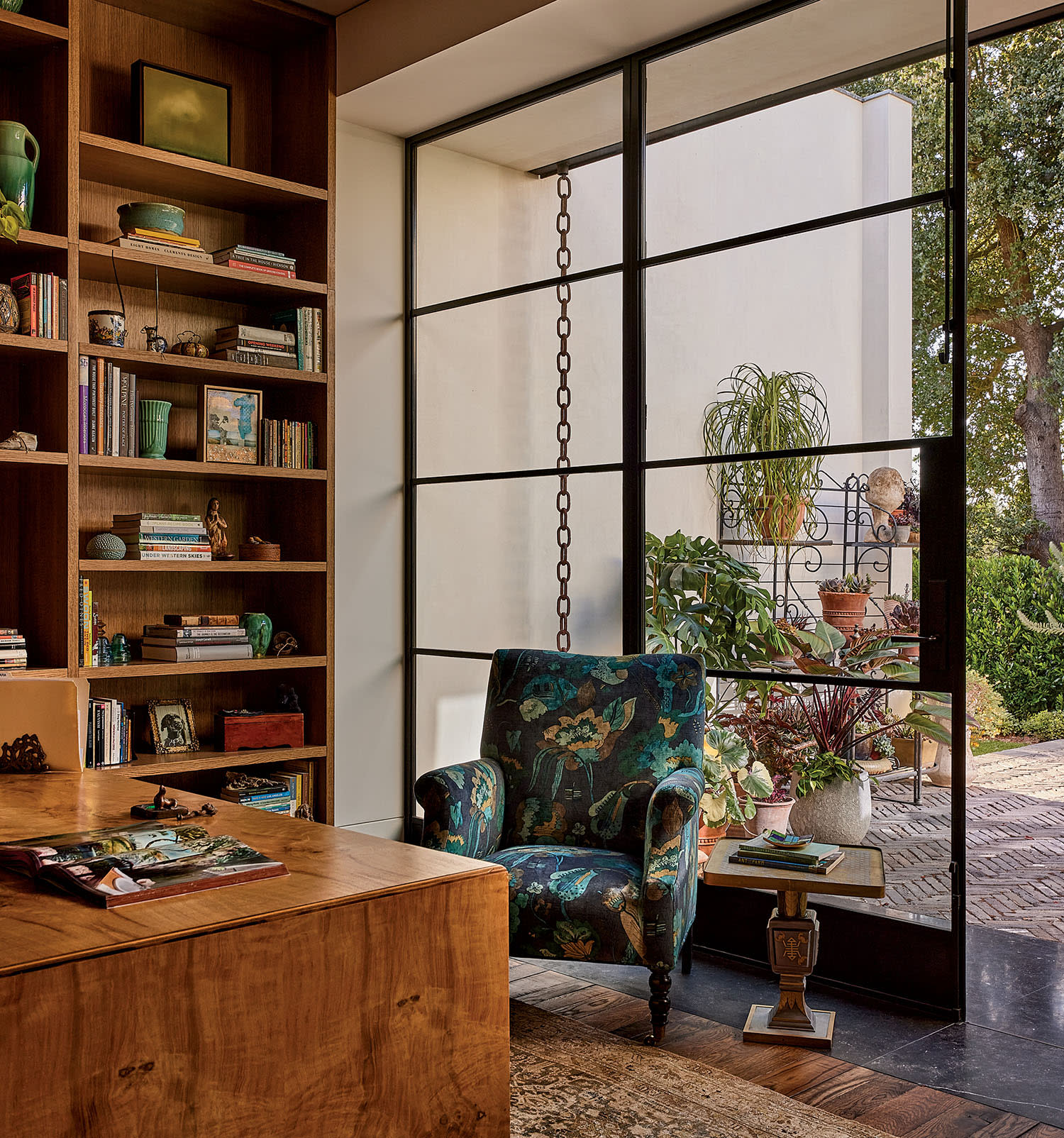 Cozy room with wooden bookshelf, colorful armchair, and large window overlooking a lush garden. Warm tones and natural light create a serene atmosphere.