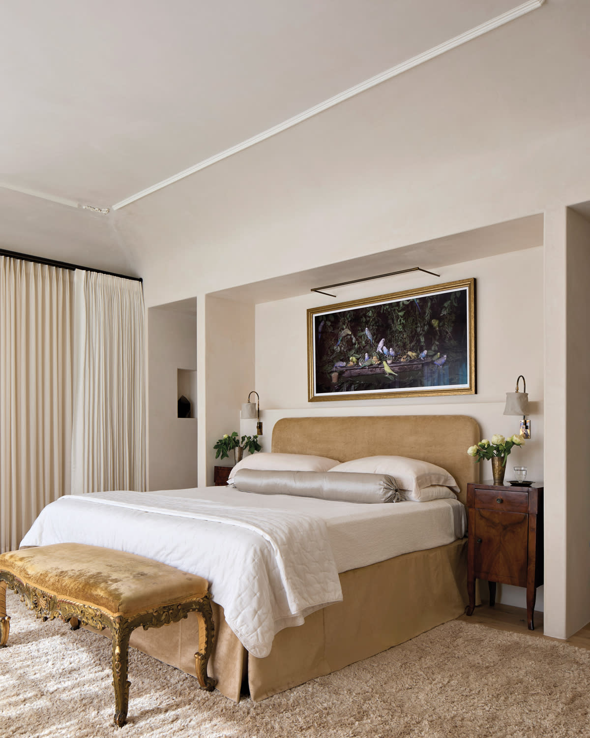 Elegant bedroom with a plush bed, gold bench, and bedside tables. A framed painting hangs above, adding sophistication.