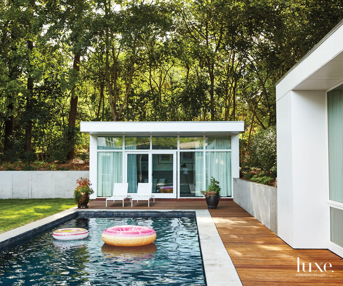 a standalone structure with a glass front sits behind a long swimming pool with wood surrounding it