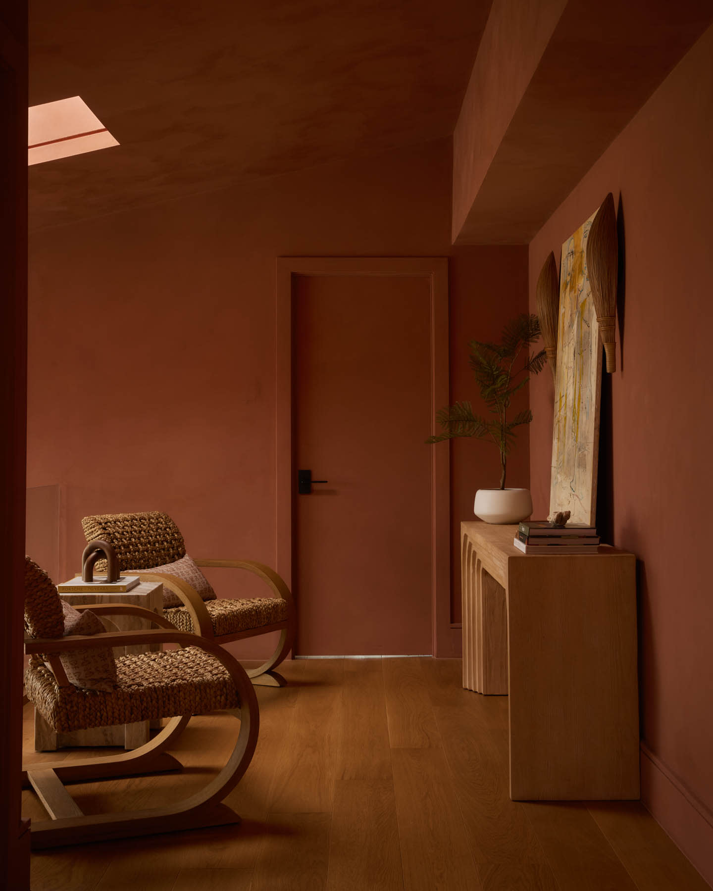 Cozy room with terracotta walls featuring natural woven chairs on light wood flooring. A minimalist console displays art, books, and a potted plant.