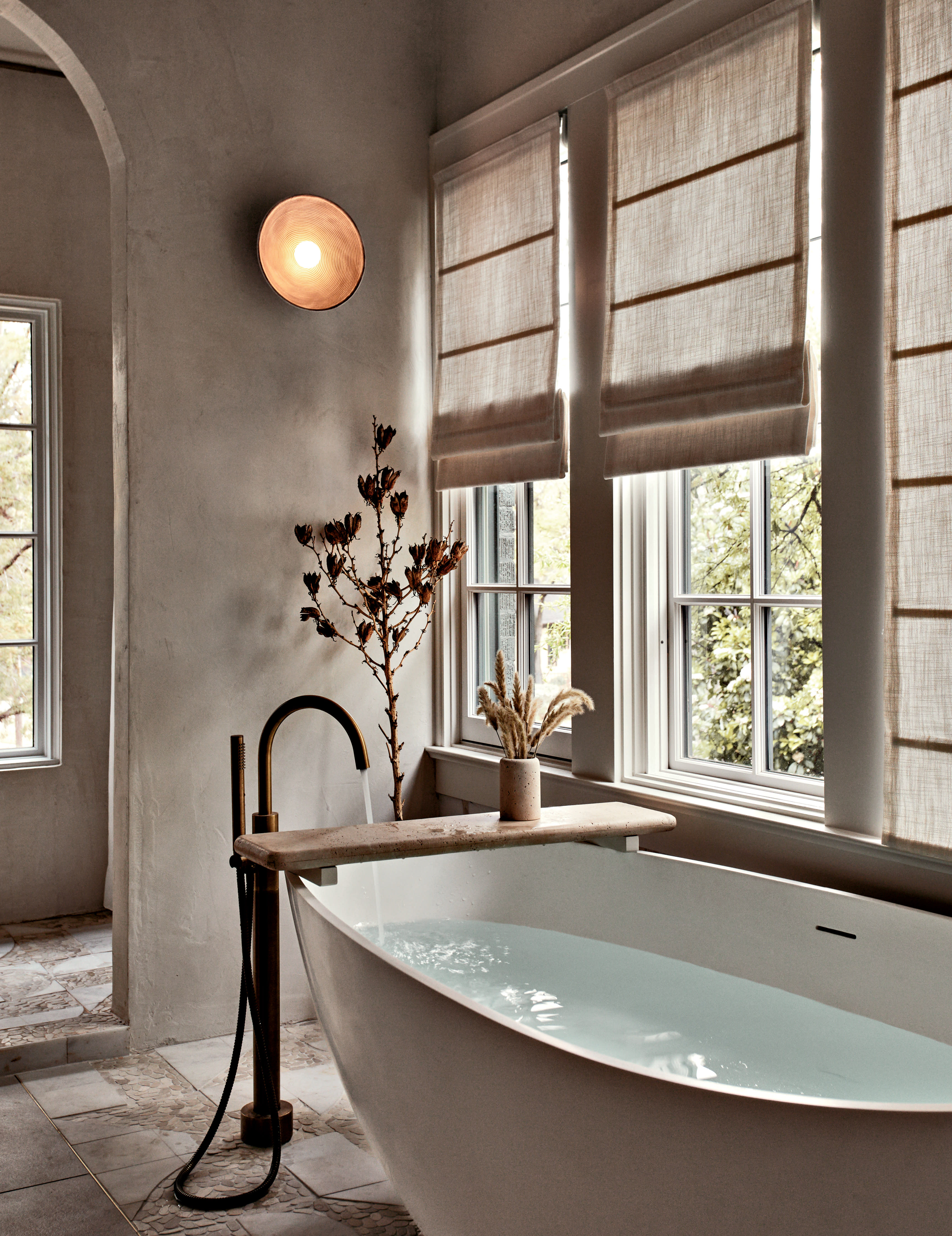 A bathroom with a freestanding stone tub and microcement walls, a round sconce light, and windows.