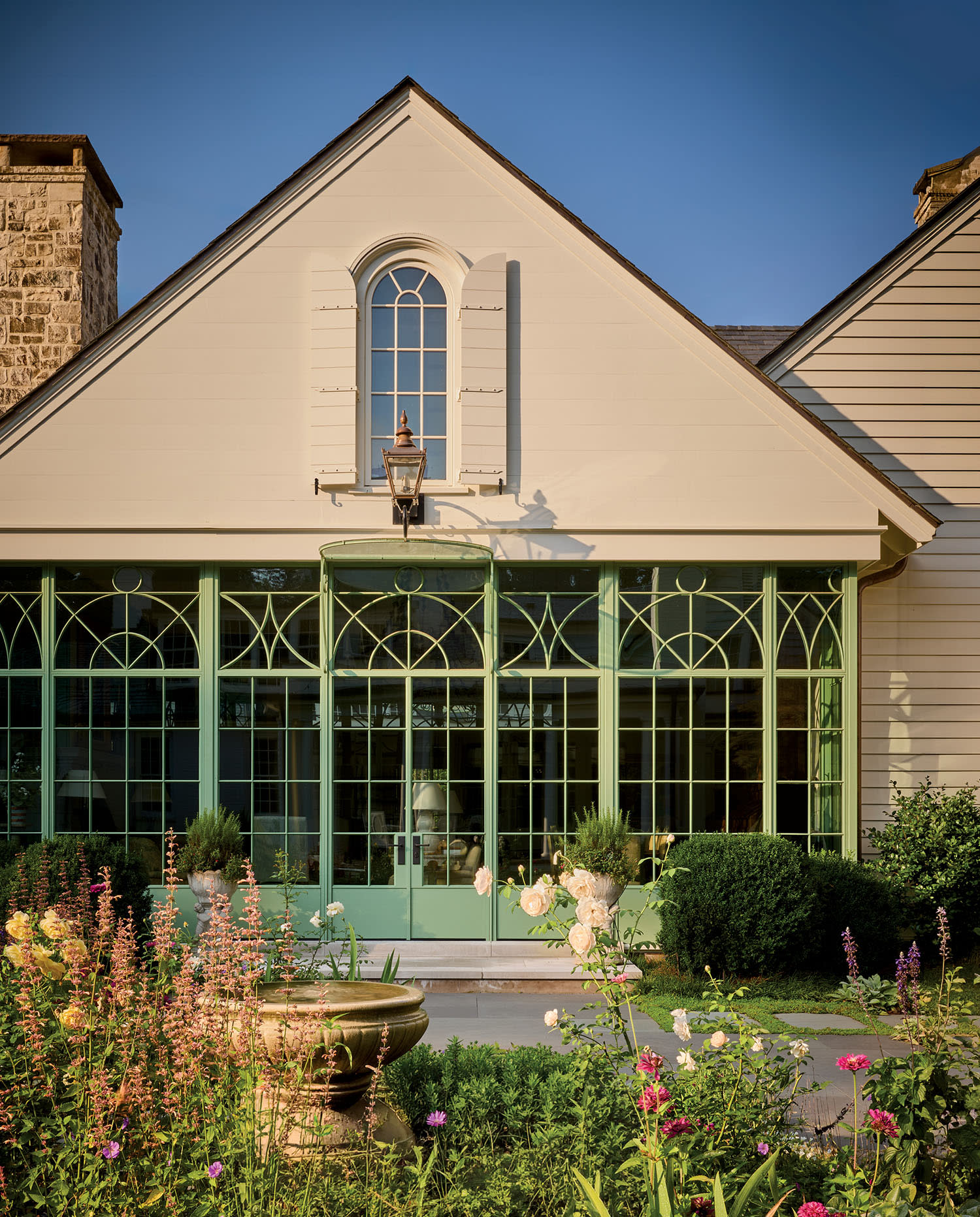 A charming house with arched windows framed in teal, surrounded by a lush garden with colorful flowers and a stone birdbath under a sunny sky.