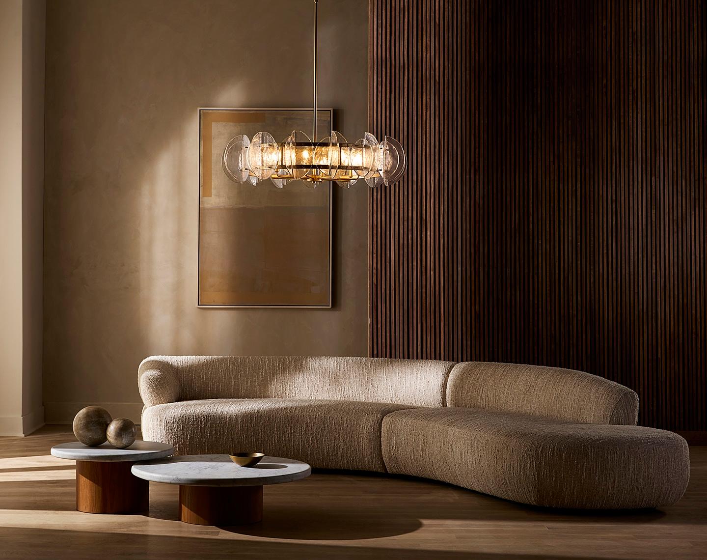 A modern living room with a curved beige sofa, two round tables, a warm chandelier, abstract artwork, and wooden accent wall, creating a cozy, elegant ambiance.