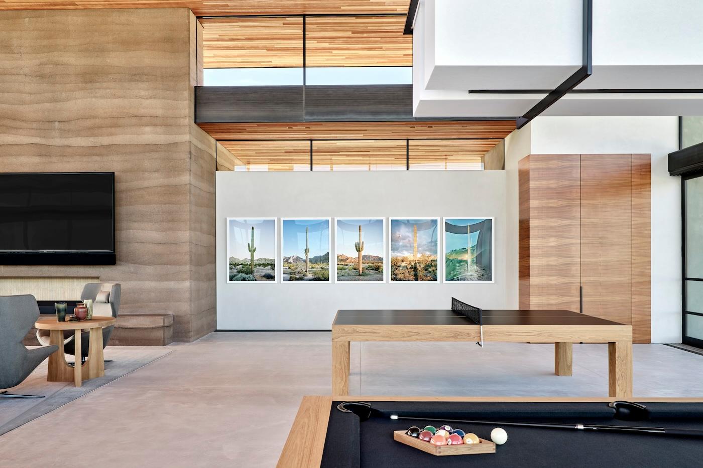 Modern living room with high ceilings, wooden accents, and large windows. Features a pool table and ping pong table, plus cactus artwork on the wall.