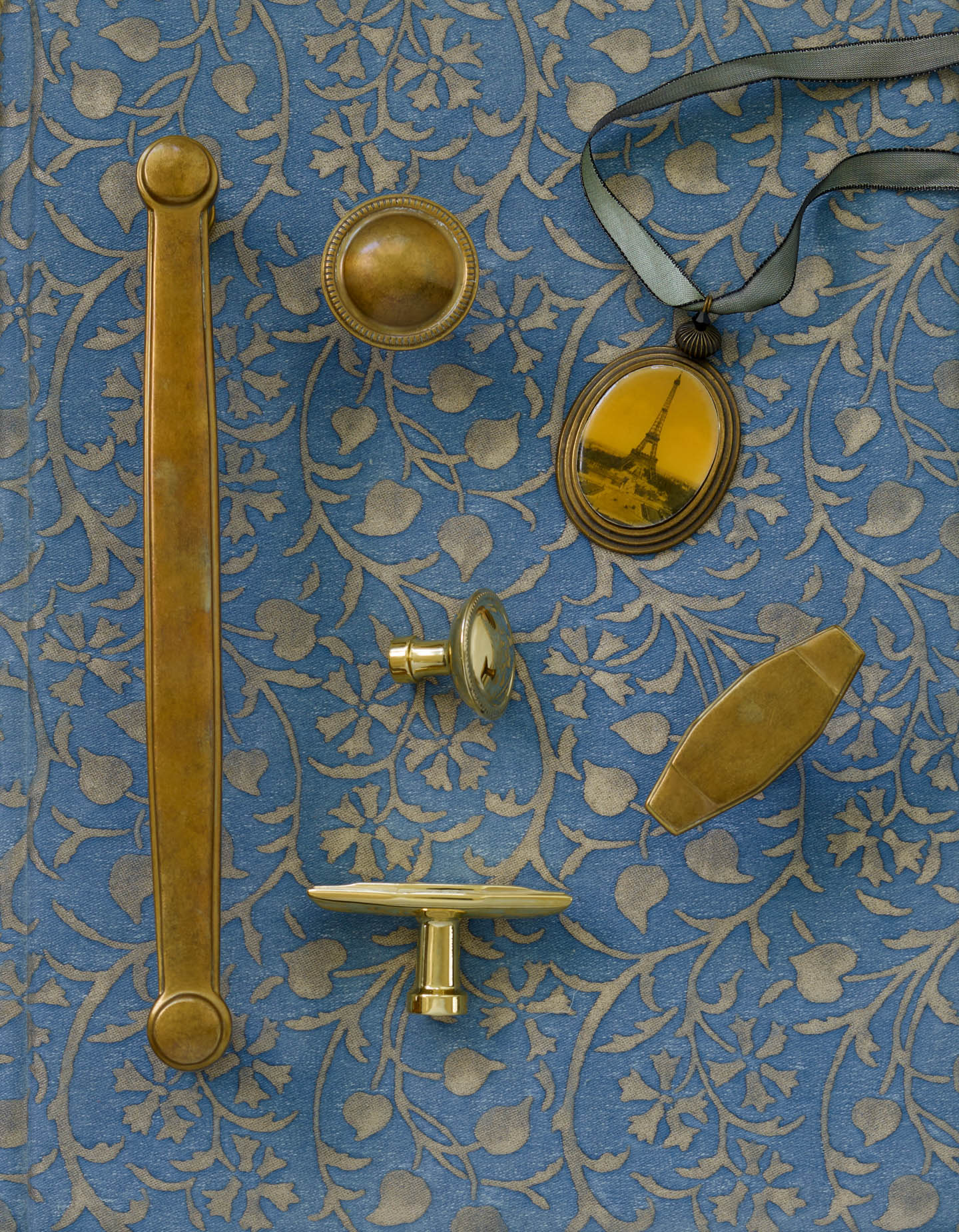 Brass door handles and knobs are arranged on embossed blue floral fabric. A pendant with an Eiffel Tower image adds a nostalgic touch.