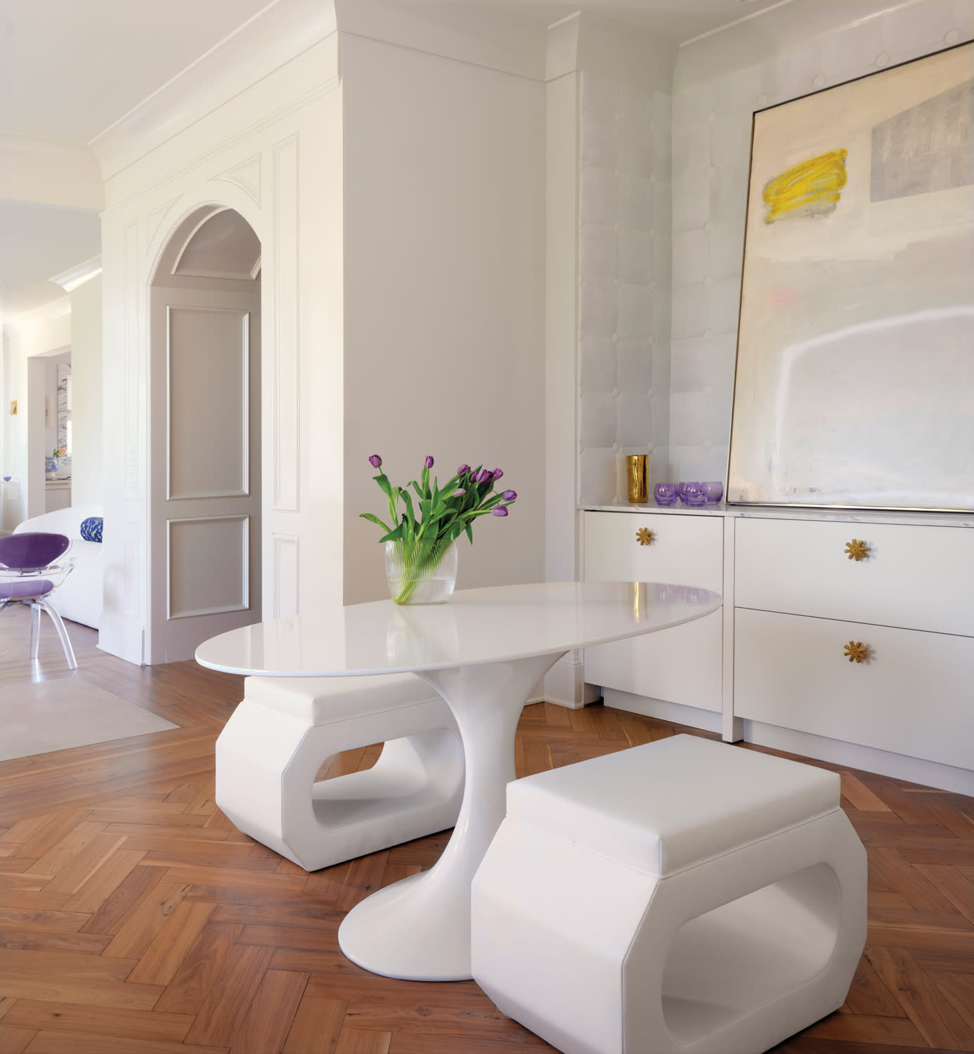 Modern interior with white furniture, featuring a sleek oval table, two geometric stools, a vase of purple tulips, and abstract art on a dresser.