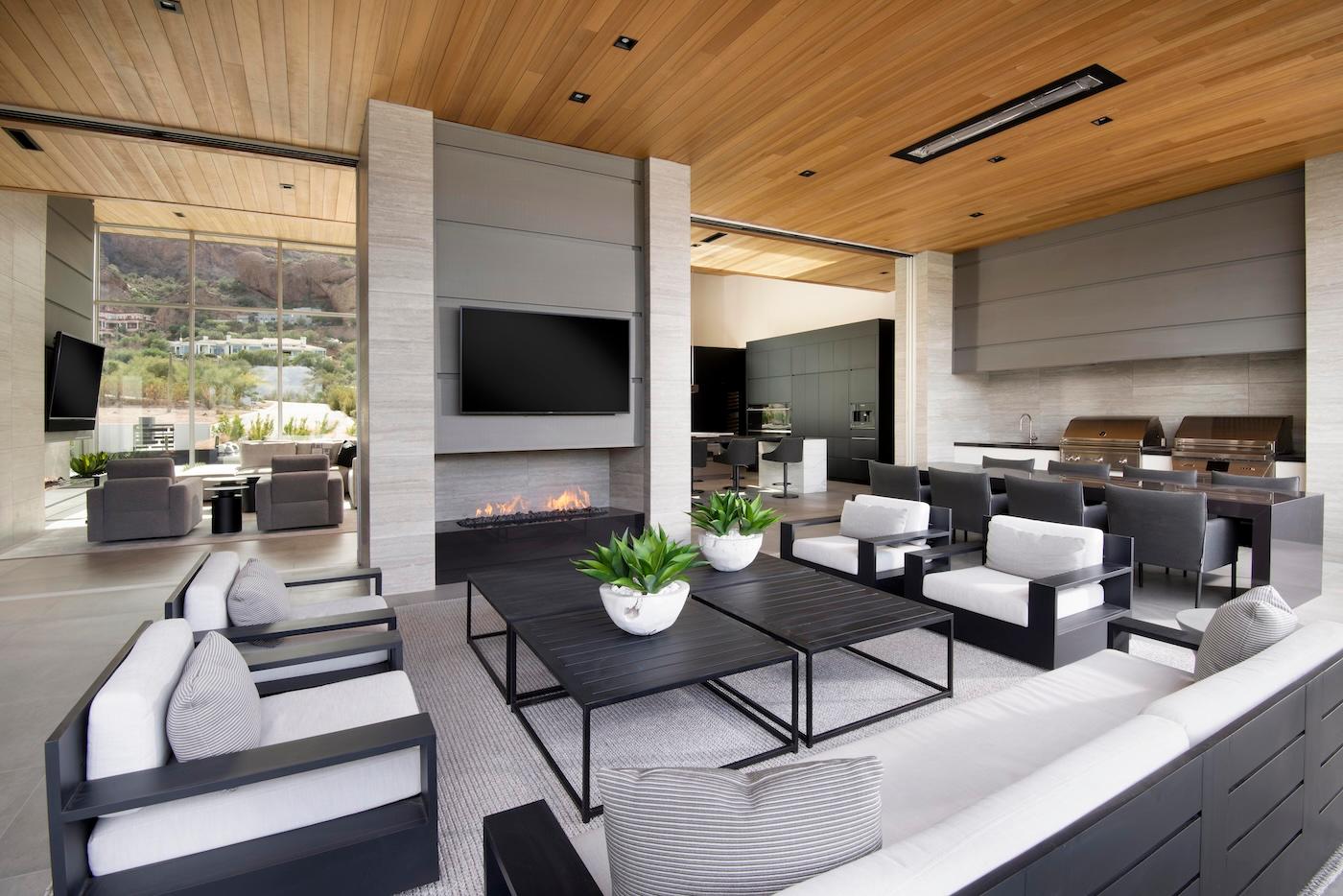 Modern open-concept living room with sleek black and white furniture, a large TV above a fireplace, and ceiling-to-floor windows showing mountain views.