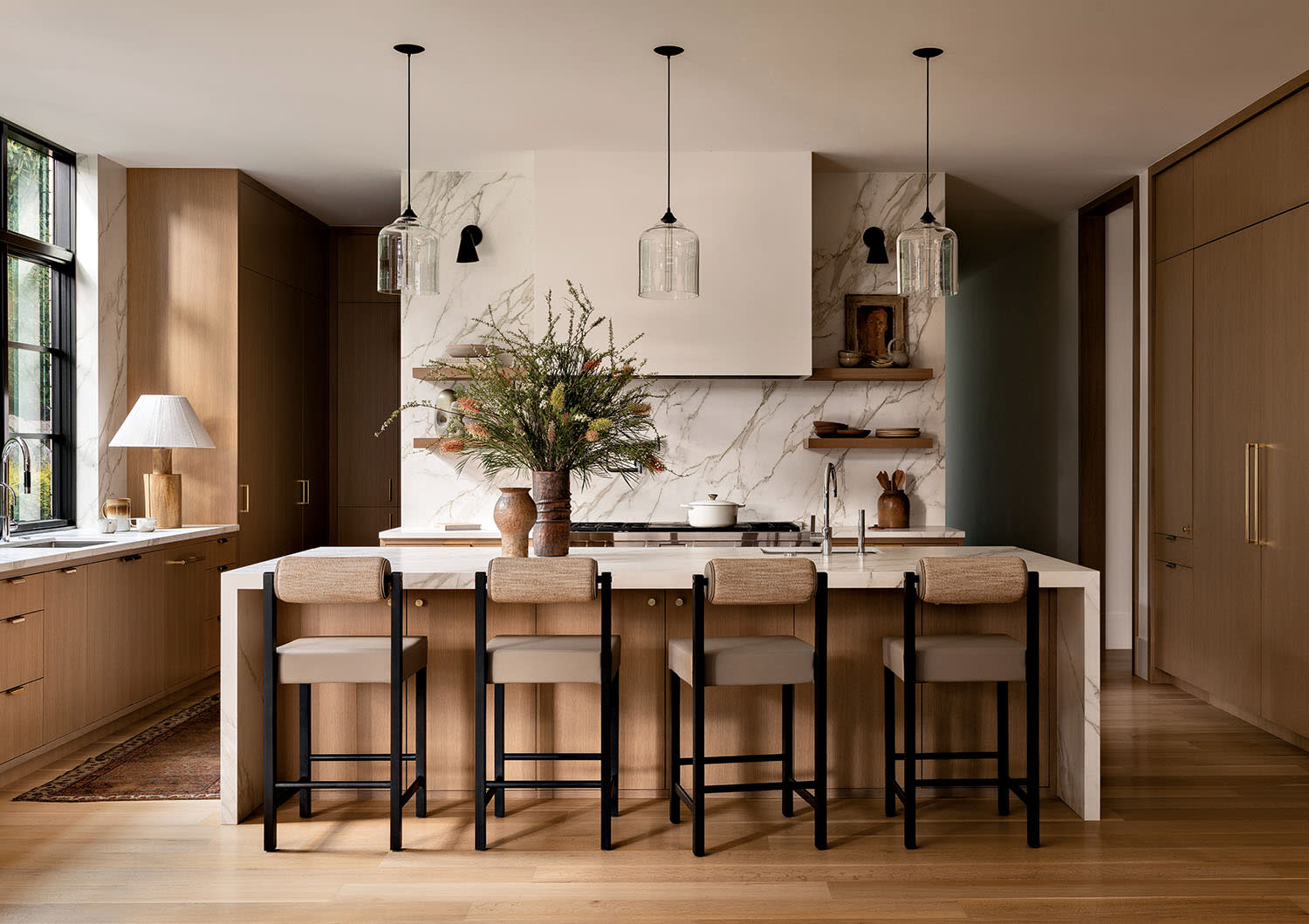 Modern kitchen with a marble island, four wooden bar stools, and three pendant lights. Light wood cabinets, open shelves, and green plant decor create a cozy, elegant feel.