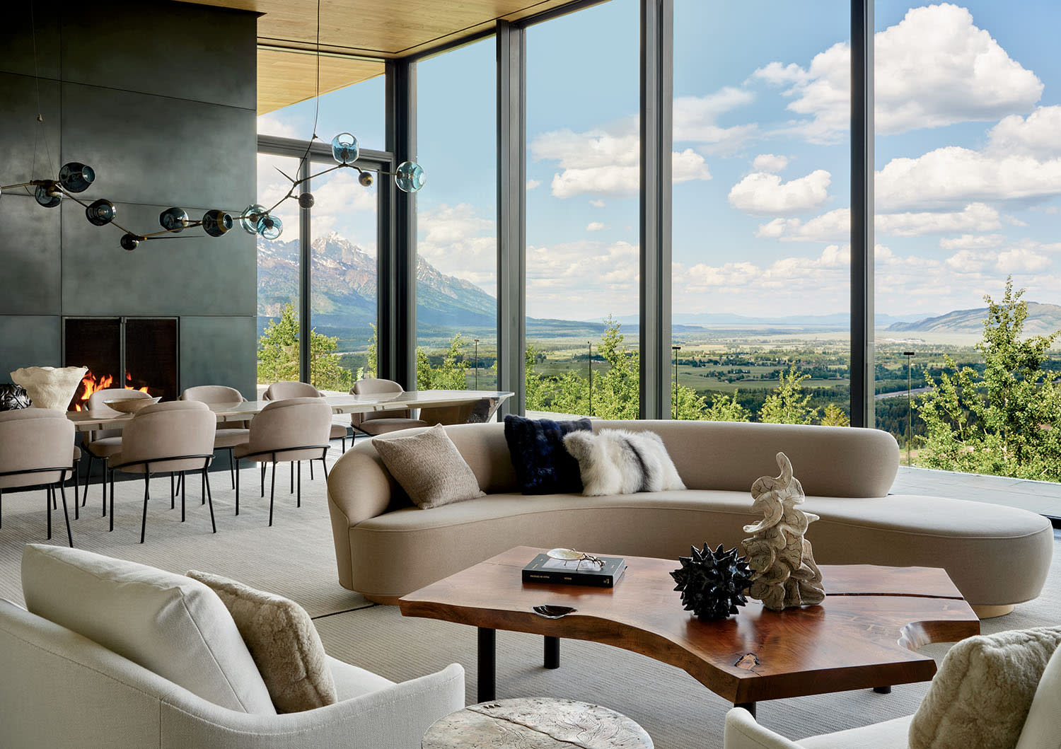 Modern living room with large windows revealing a scenic mountain view. Features beige decor, a cozy fireplace, and a wooden coffee table adding warmth.