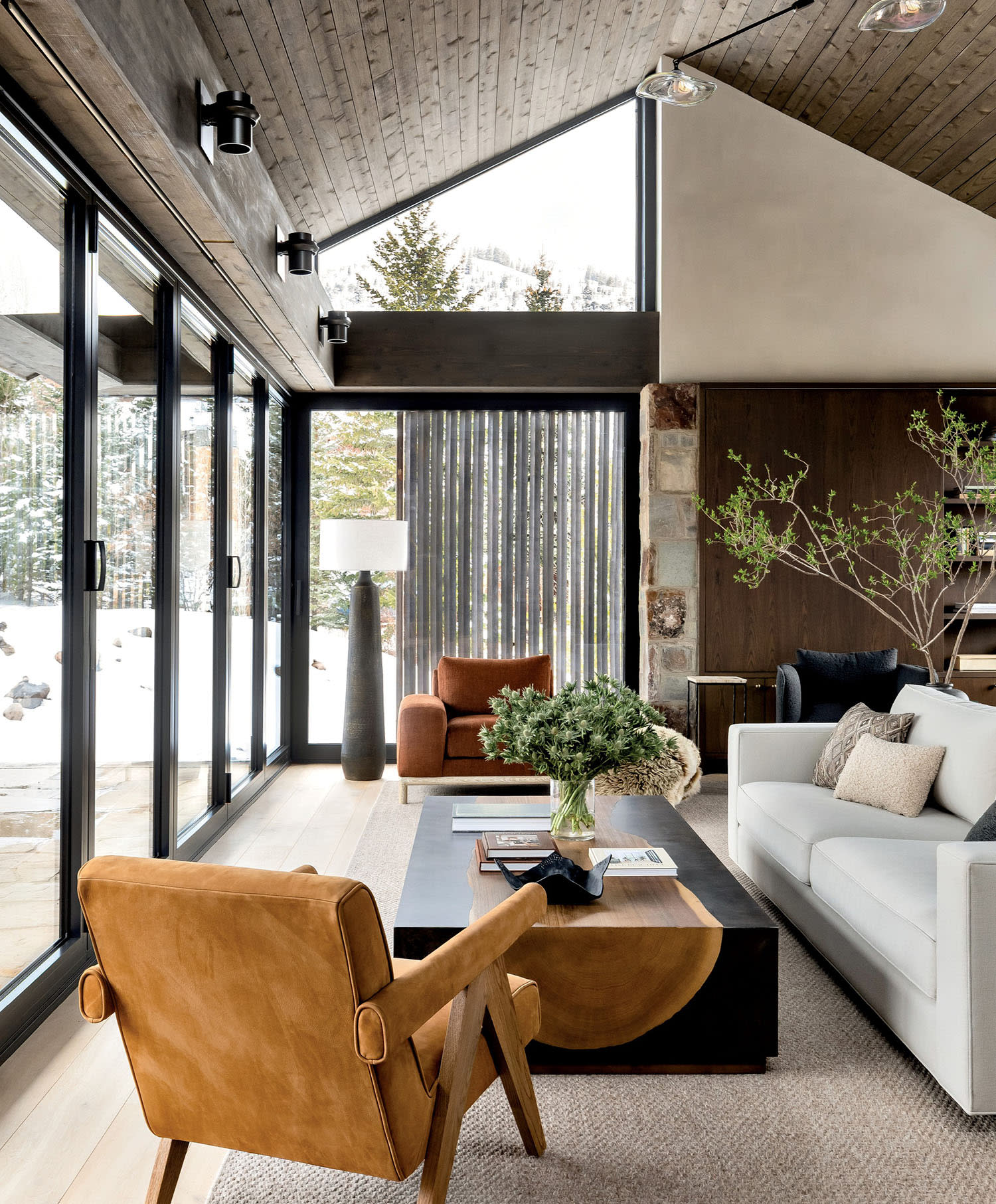 Modern living room with large floor-to-ceiling windows, warm wood tones, leather chairs, a white sofa, and greenery on a chic black coffee table. Cozy and inviting.