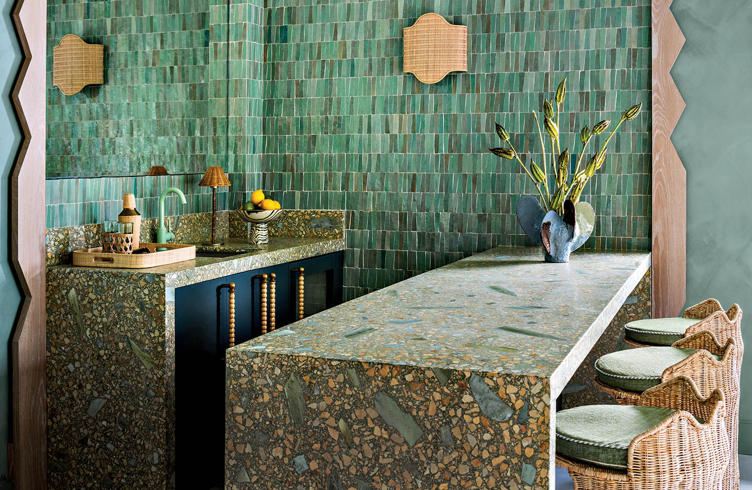 A stylish bar with green tiled walls and a terrazzo countertop. Features wicker stools, a vase with tall greenery, and ambient lighting, creating a cozy ambiance.