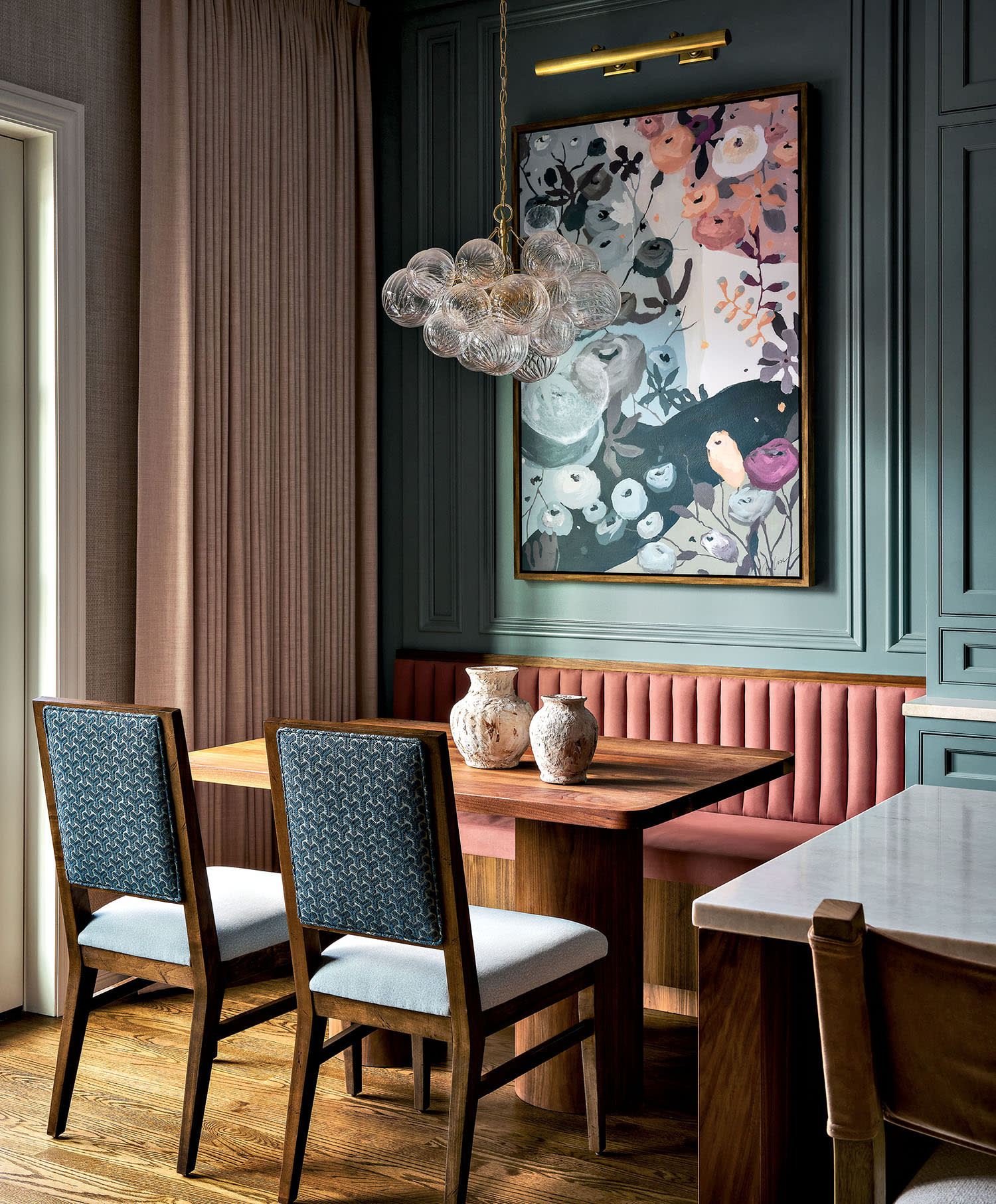 A small dining nook with a banquette and two arm chairs set beneath a bubble chandelier and artwork.