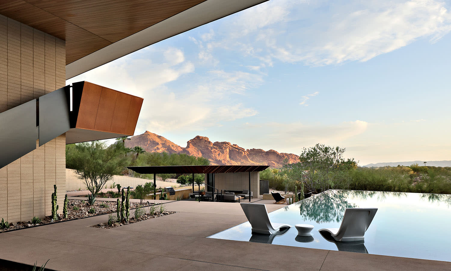 Modern desert home with angular architecture, featuring a sleek patio and reflective pool. Distant red rocky mountains under a blue sky evoke serenity.