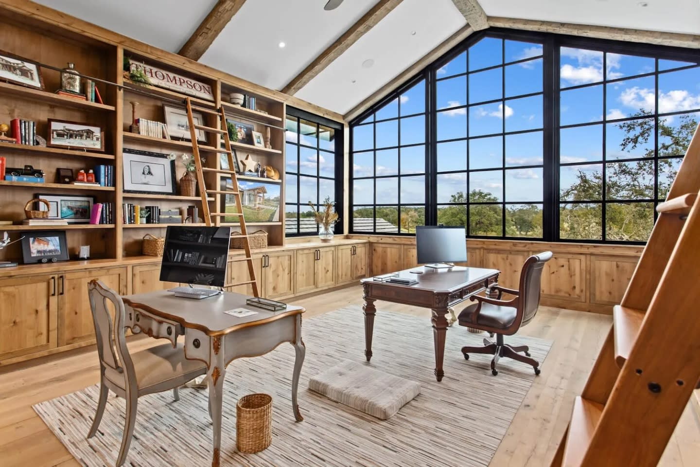 Spacious home office with wooden bookshelves, a ladder, and large windows offering a scenic view. A desk with a computer and chair sits on a light rug.