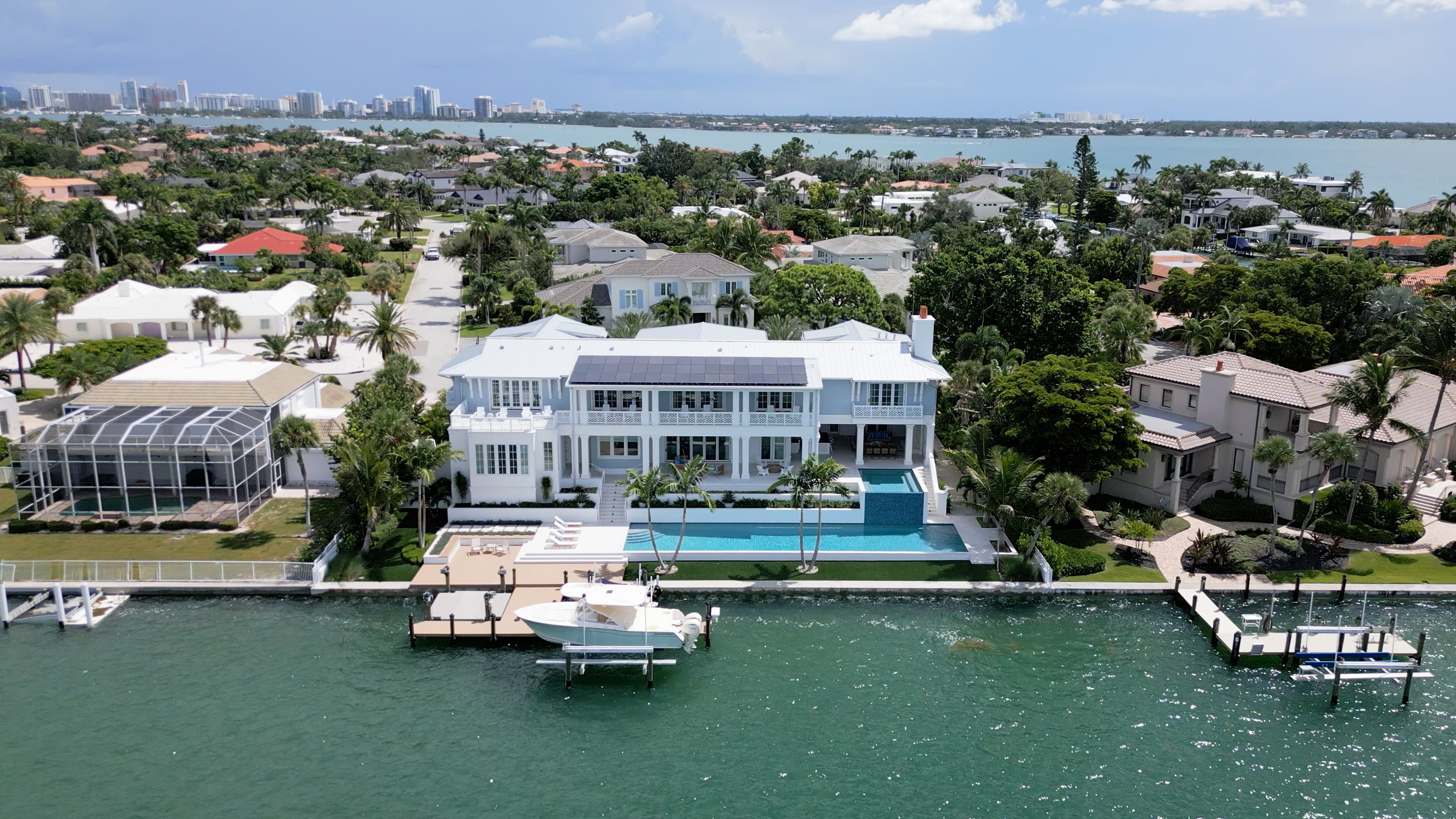 Aerial view of a luxurious waterfront mansion with a white facade, surrounded by lush greenery and neighboring homes.