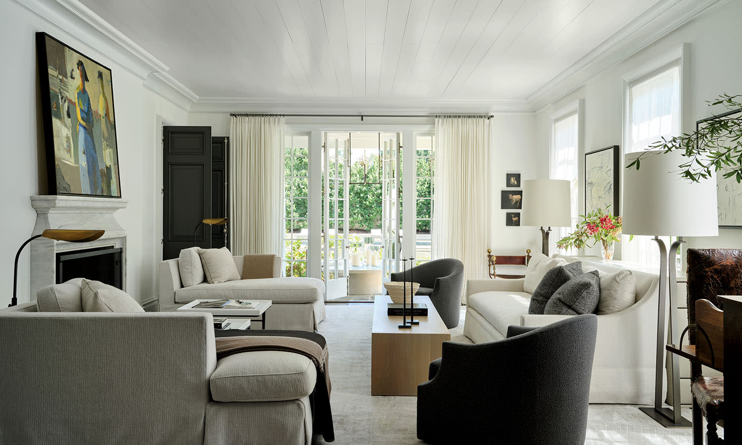 Elegant living room with neutral tones, featuring plush sofas, modern art above a fireplace, and large windows with sheer curtains overlooking a garden.