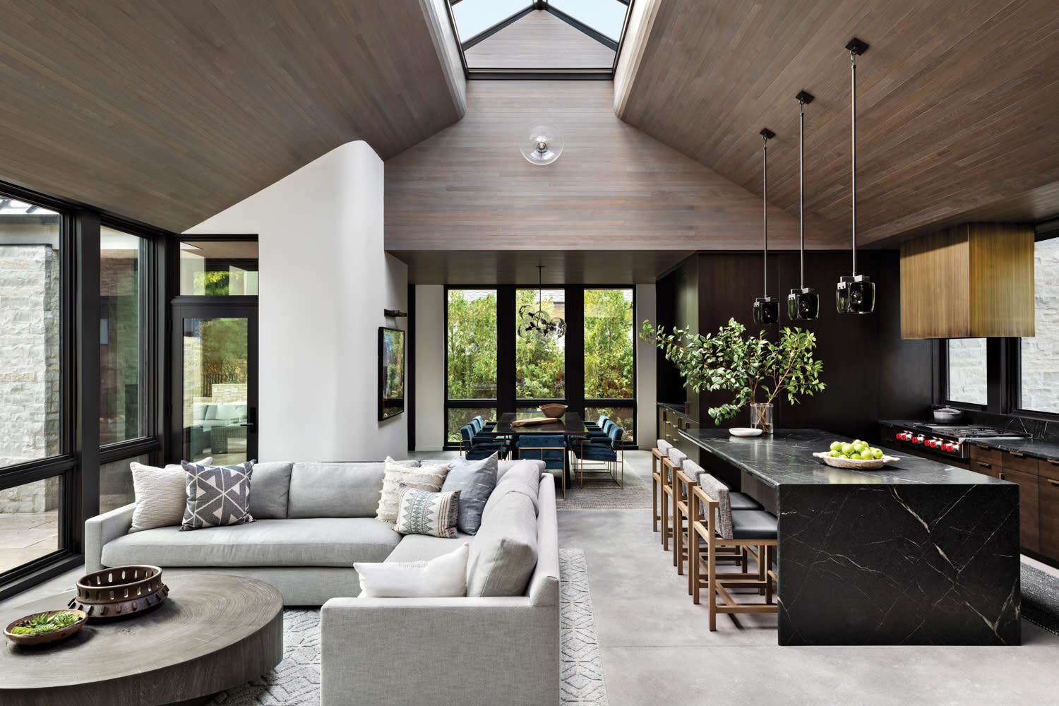 room with a gray sectional, floor-to-ceiling windows, kitchen island and glass pendants by En Masse