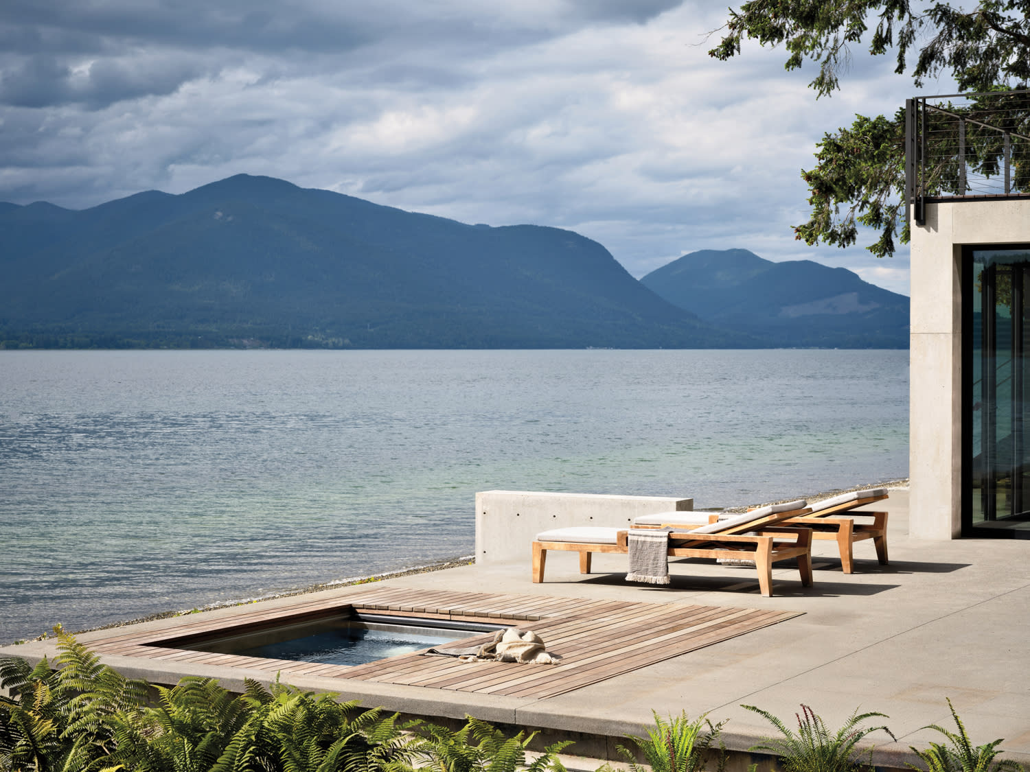 Modern lakeside patio with two wooden lounge chairs and a hot tub. Overlooks calm water and distant mountains under a cloudy sky, conveying tranquility.