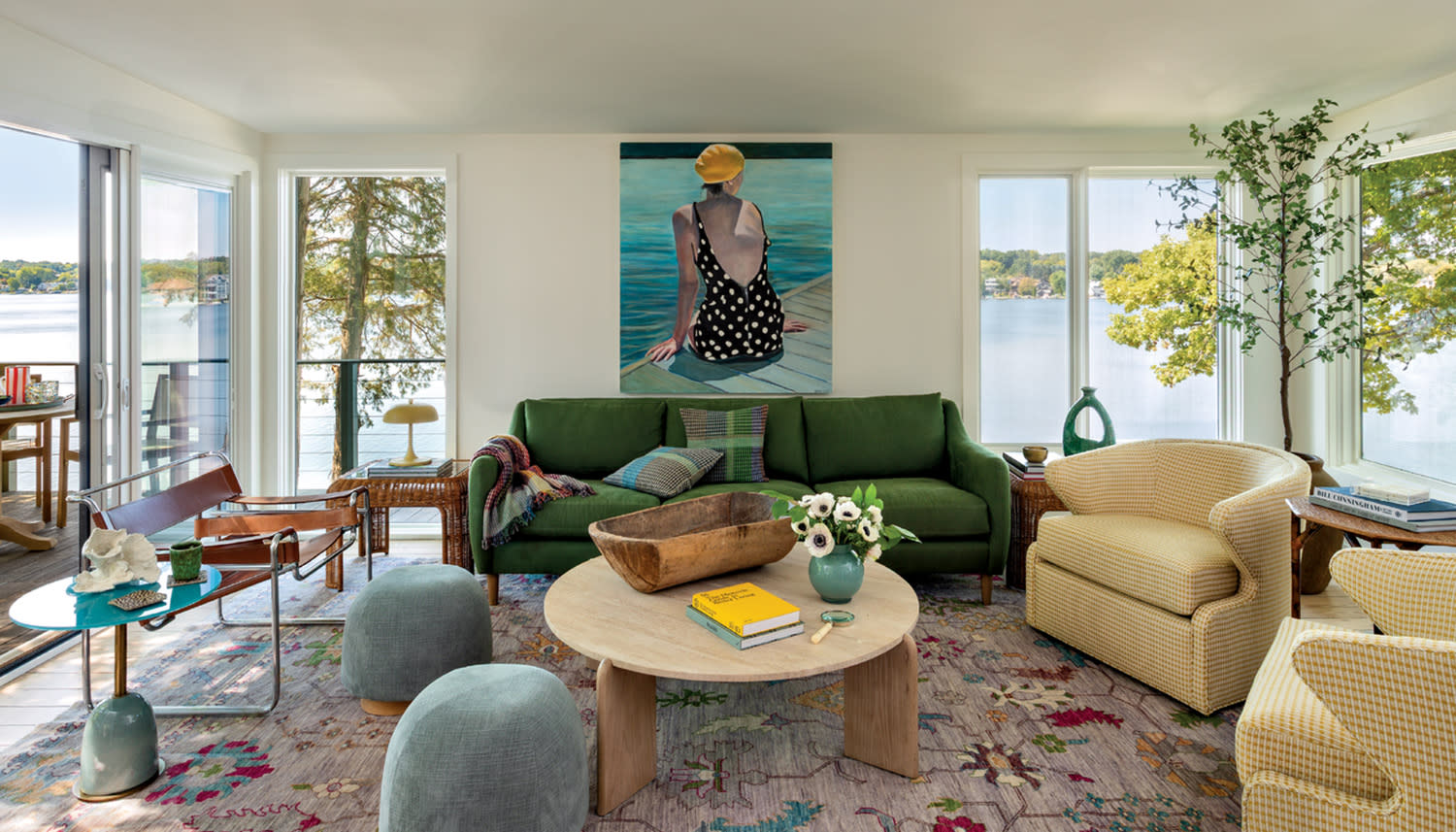 Bright living room with lake view, featuring green sofa, abstract rug, large painting, circular wood table, and plants. Relaxed, inviting atmosphere.