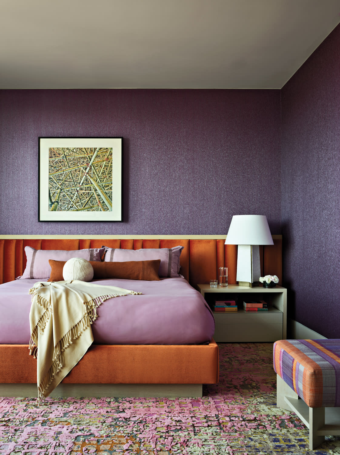A modern bedroom features a purple accent wall, an orange bed with purple bedding, and a beige throw. A bedside table holds a white lamp. Warm, cozy ambiance.