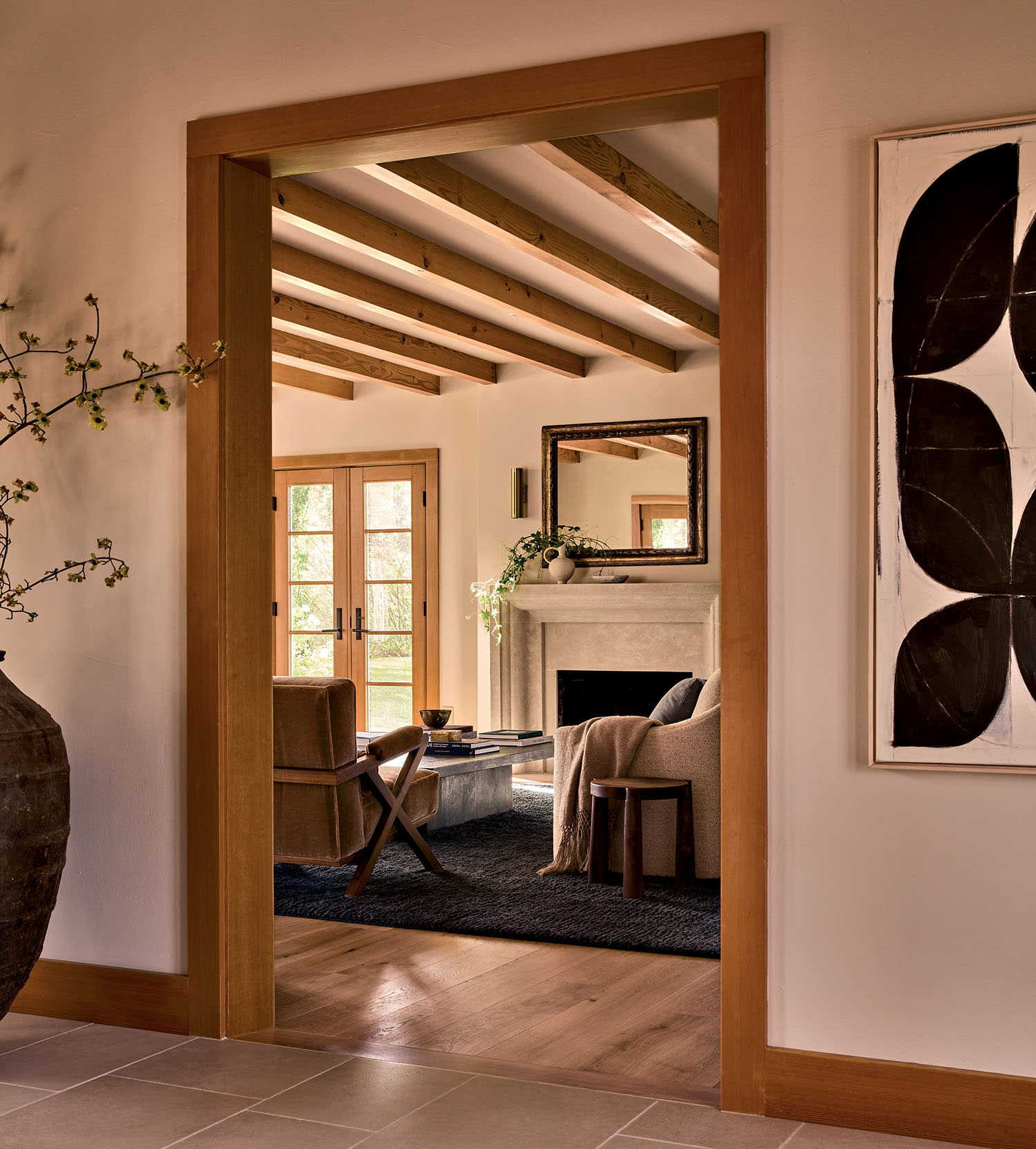 Cozy living room with wooden beams, a stone fireplace, and a large mirror. Warm tones, soft armchair, and abstract art create an inviting atmosphere.