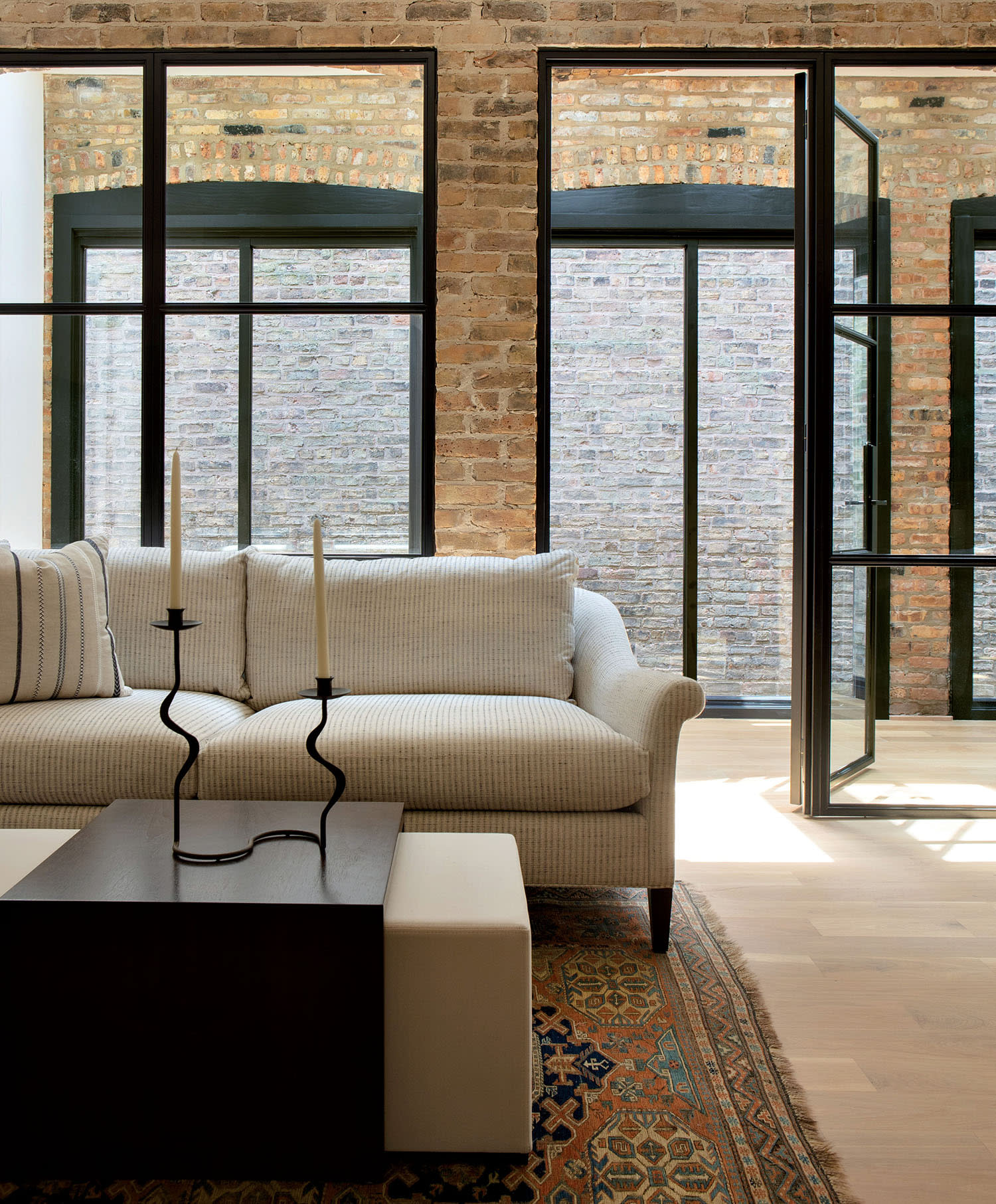 A gray couch sits in front of a coffee table decorated with candlesticks, and glass and brick walls behind it.