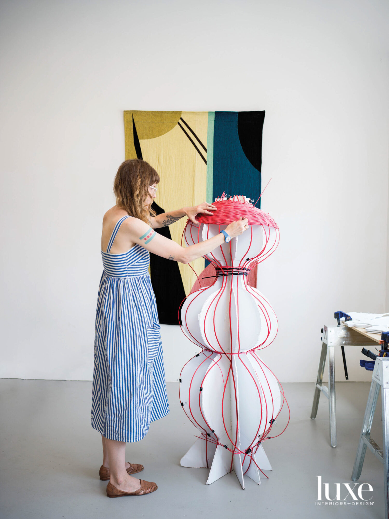 The artist weaving a large pink basket. A textile hangs on the wall.