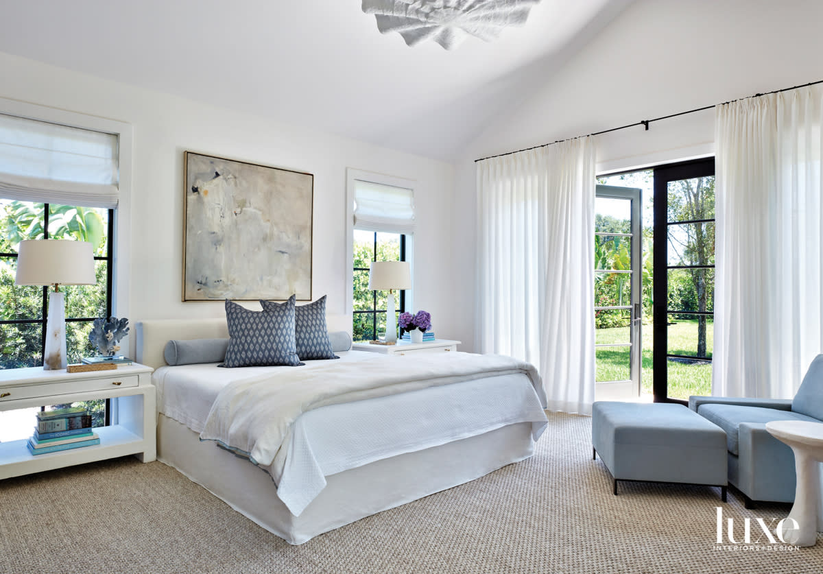 primary bedroom with white walls, bed and draperies and blue armchair, ottoman and pillows