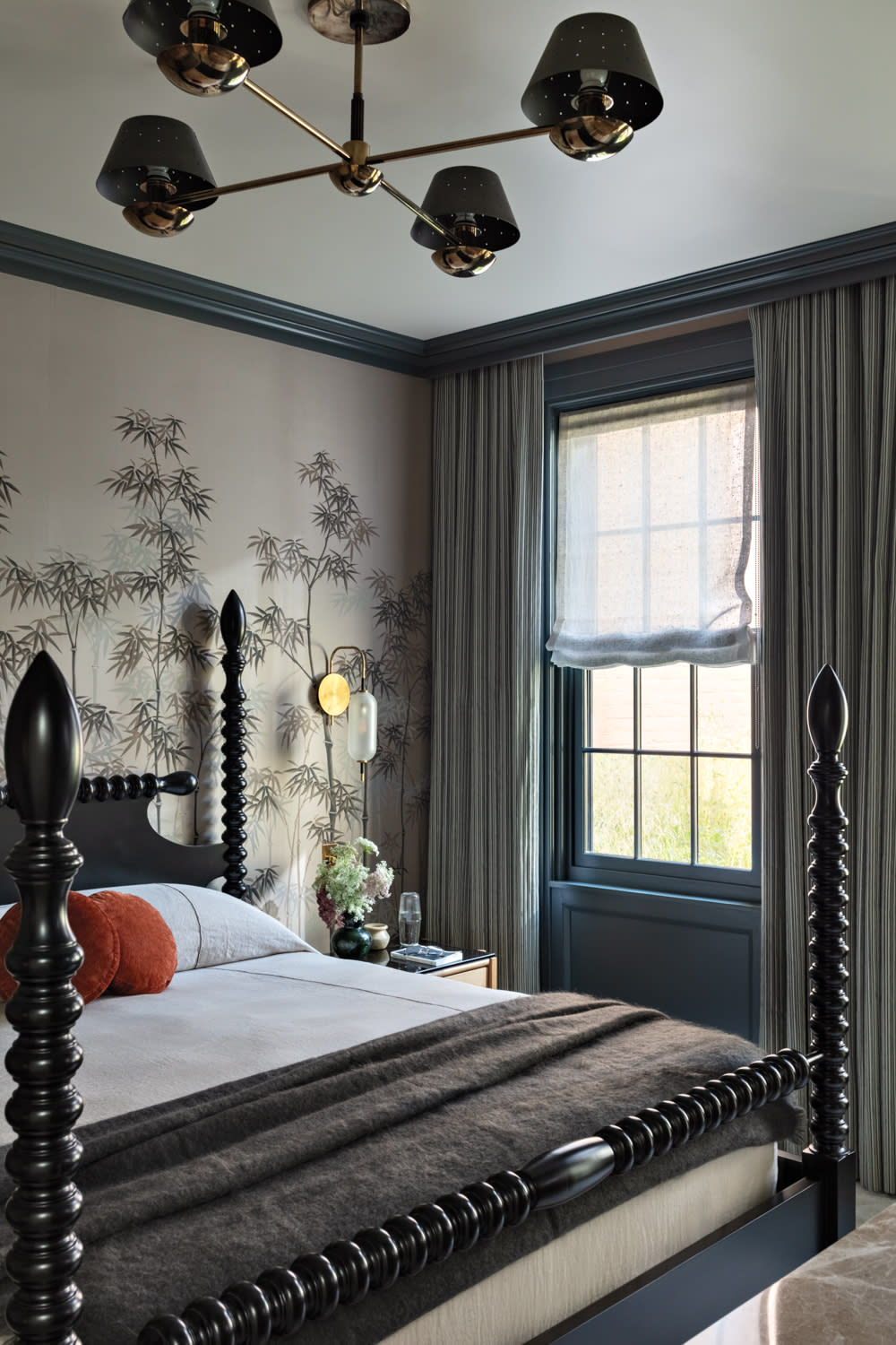Sophisticated bedroom featuring a dark spindle bed with cozy bedding, bamboo-patterned wallpaper, elegant chandelier, and soft, filtered light from a window.