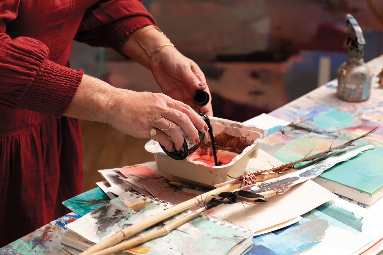 Two hands pouring paint into a container on top of a table covered in paintings.