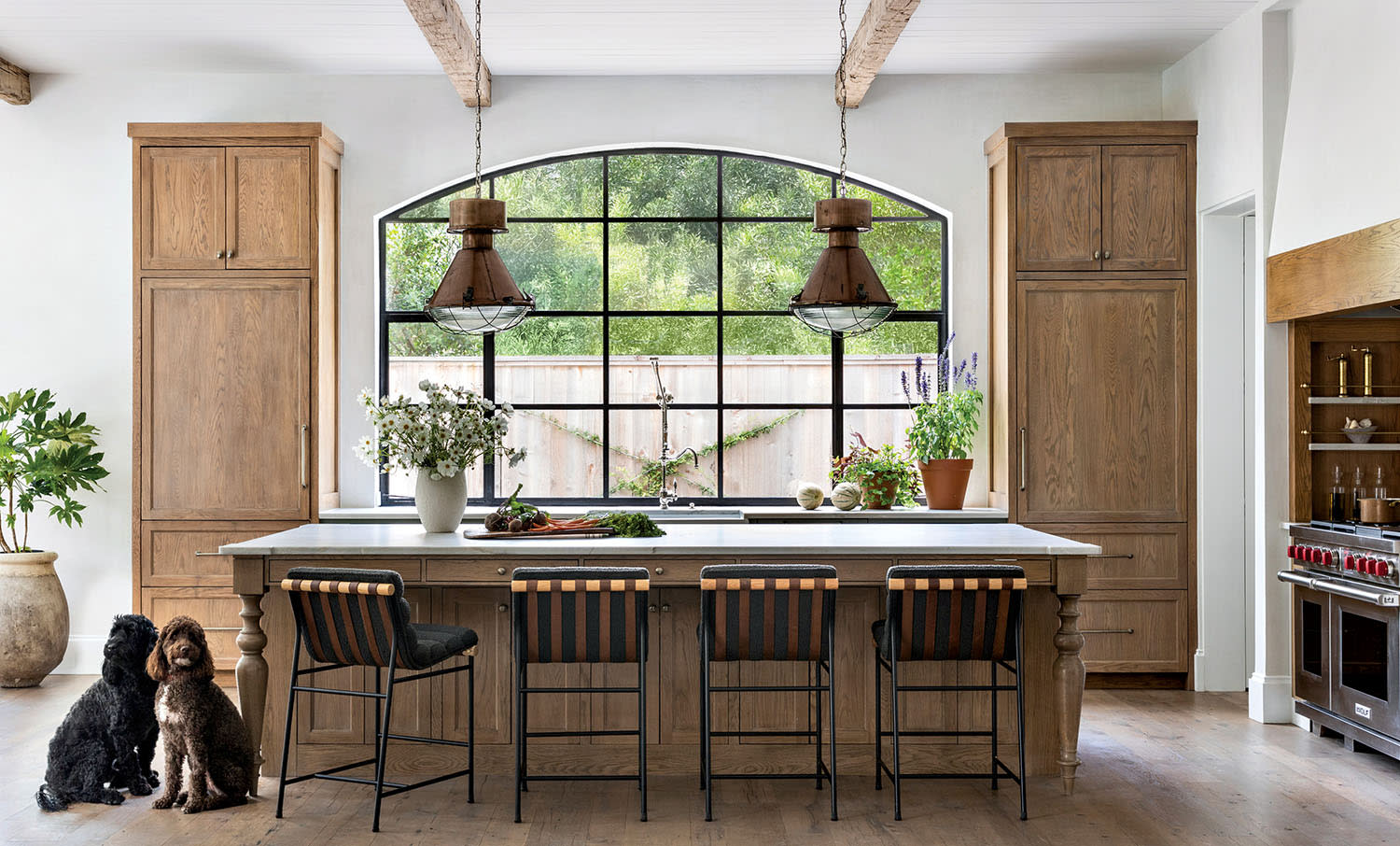 Spacious kitchen with large arched window, wooden cabinets, and hanging lights. A kitchen island with stools and two dogs sit on the wooden floor.