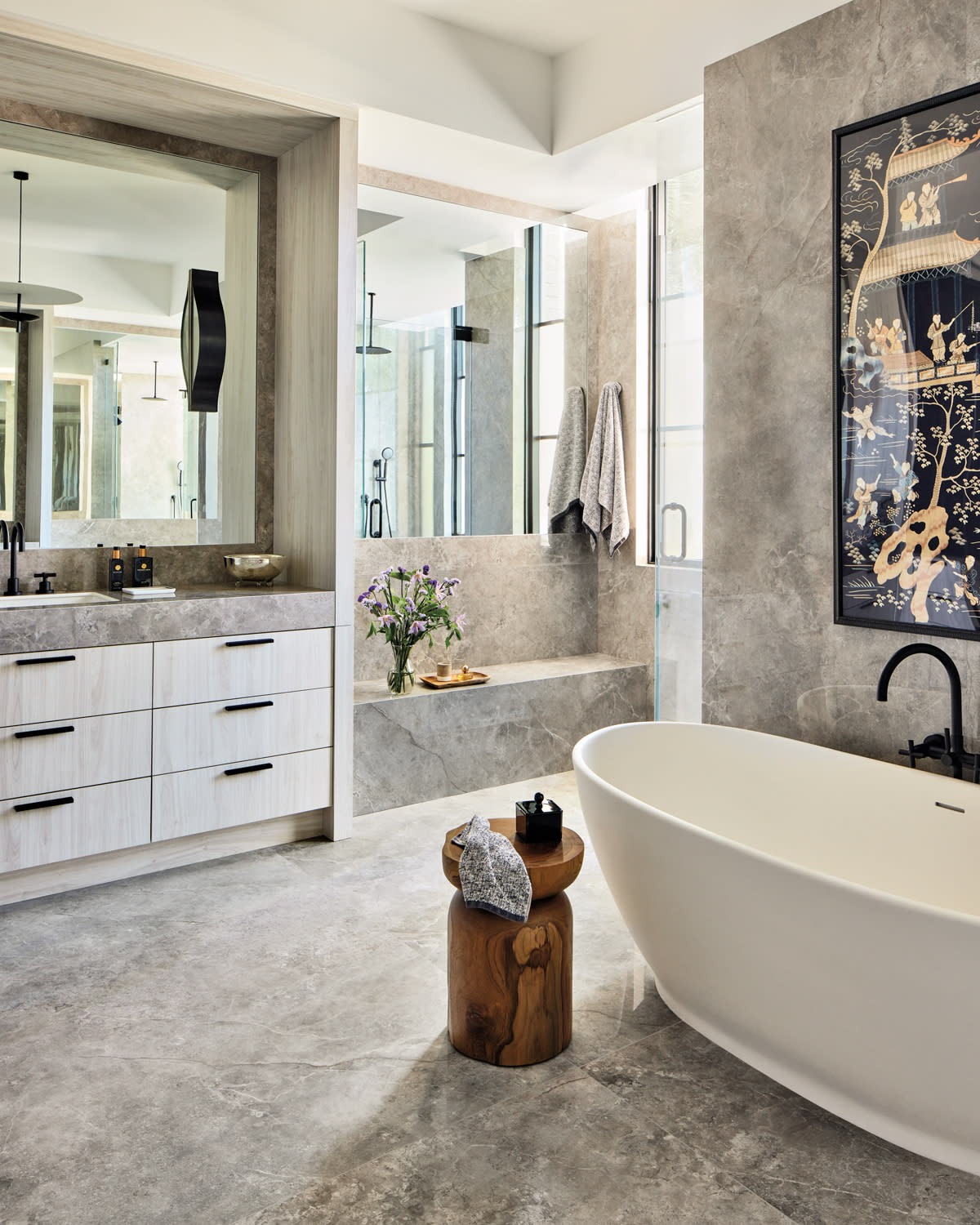 Luxurious bathroom with a freestanding white tub, black faucet, gray marble tiles, and sleek wood cabinet. Artistic wall art and flowers add elegance.