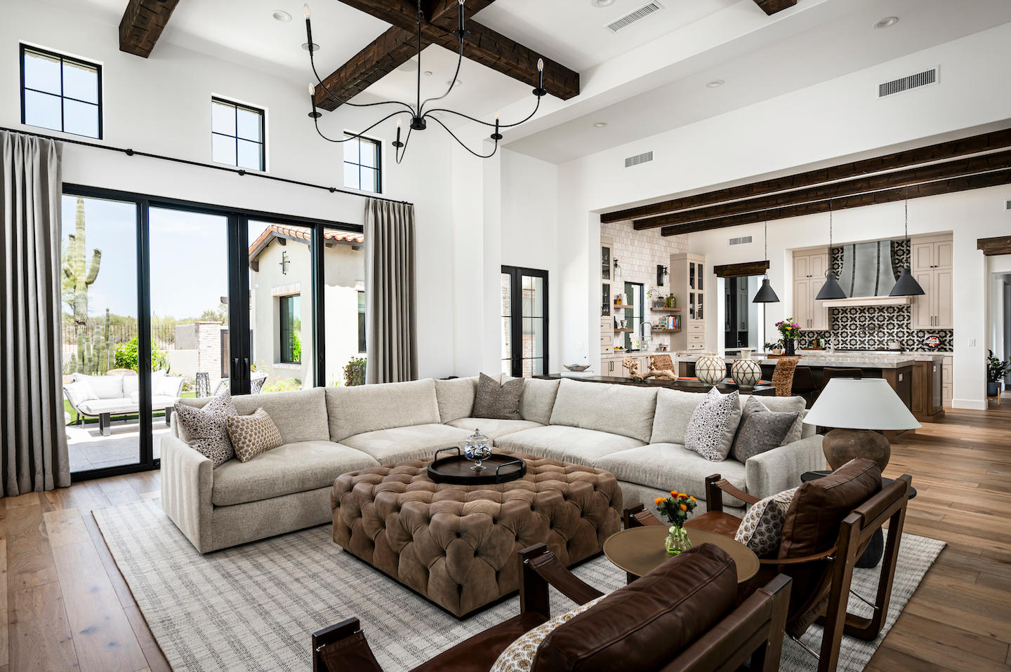 Airy great room with soaring white walls, exposed dark-wood ceiling beams, and a sculptural black chandelier over an oversized cream-linen sectional. A tufted caramel-suede ottoman centers the seating, while sliding glass doors frame desert views and invite sunlight onto wide-plank floors. In the background, the open kitchen shows patterned Spanish tile behind a stainless range hood, pendant lights, and a mix of painted and walnut cabinetry for a relaxed luxe vibe.