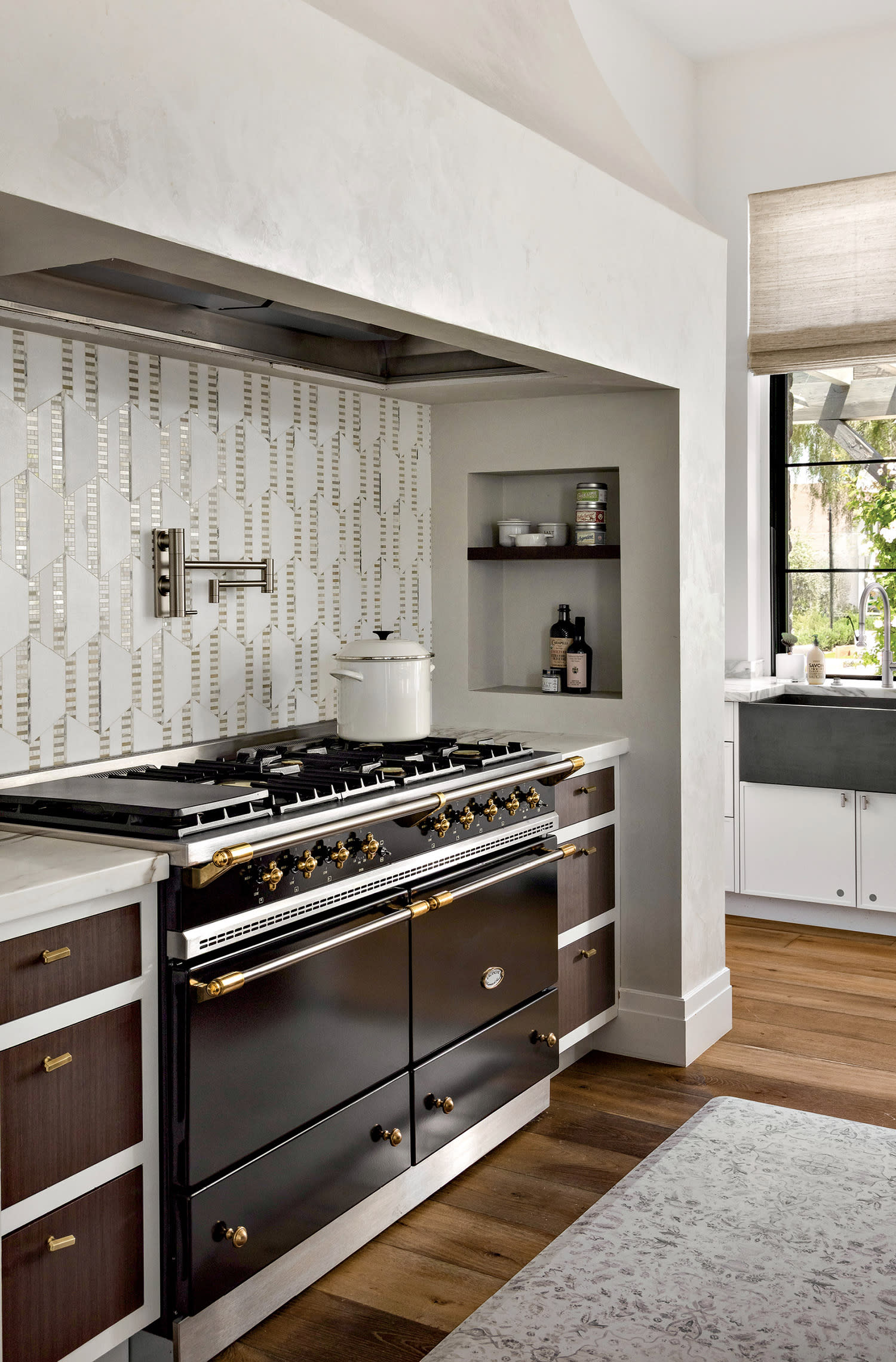 Sleek kitchen with a black stove, brass knobs, geometric tile backsplash, and light wood cabinets. Sunlit window with a view outside, modern feel.