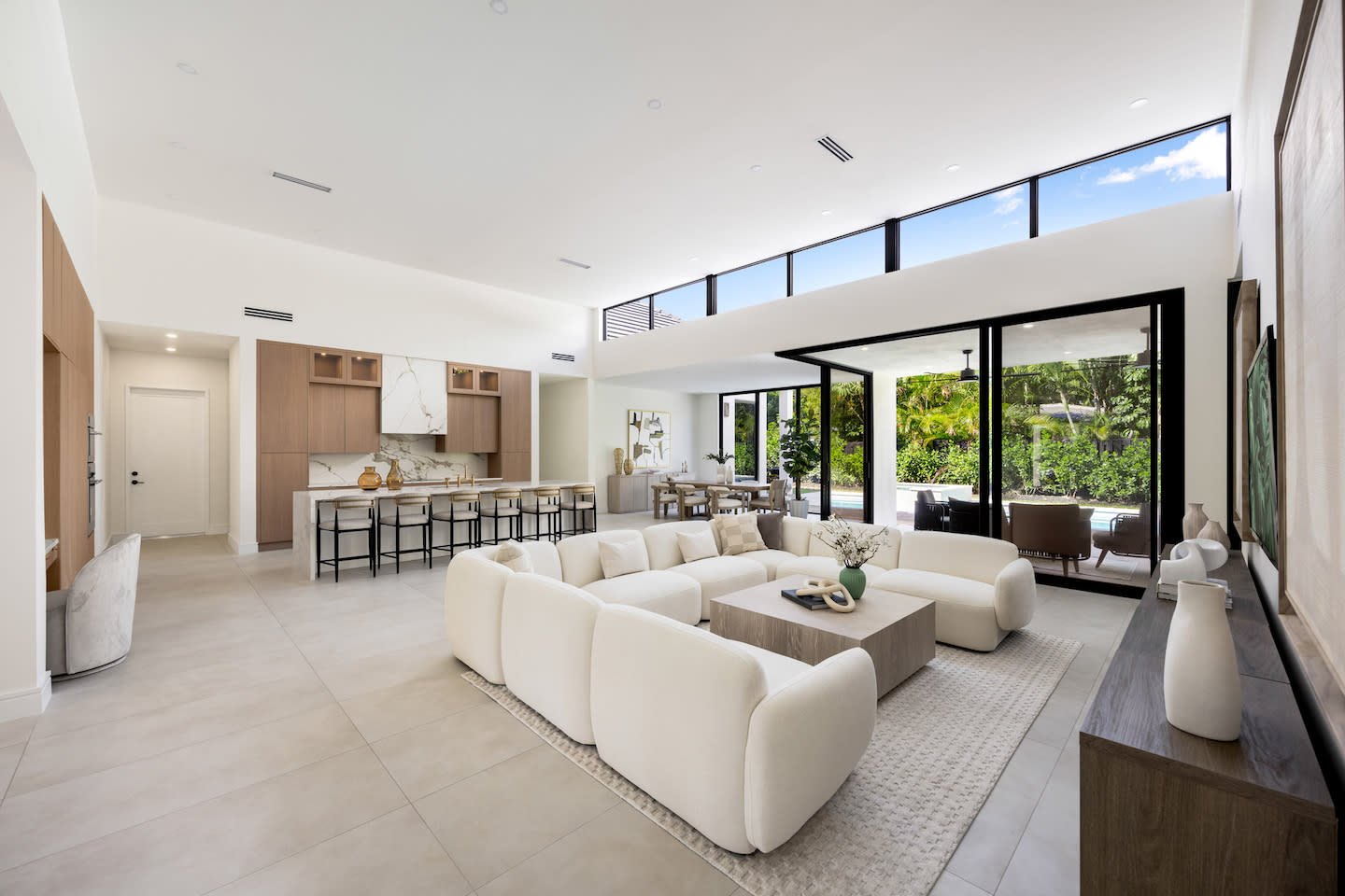Airy, open-plan living space featuring a cloud-like cream modular sofa and oak coffee table beneath a ribbon of clerestory windows that flood the room with light. A waterfall-edge marble island anchors the minimalist wood kitchen, while floor-to-ceiling sliders disappear to reveal a covered patio and pool bordered by dense tropical greenery. Neutral porcelain tiles and sculptural ceramic accents tie together the relaxed, indoor-outdoor vibe.