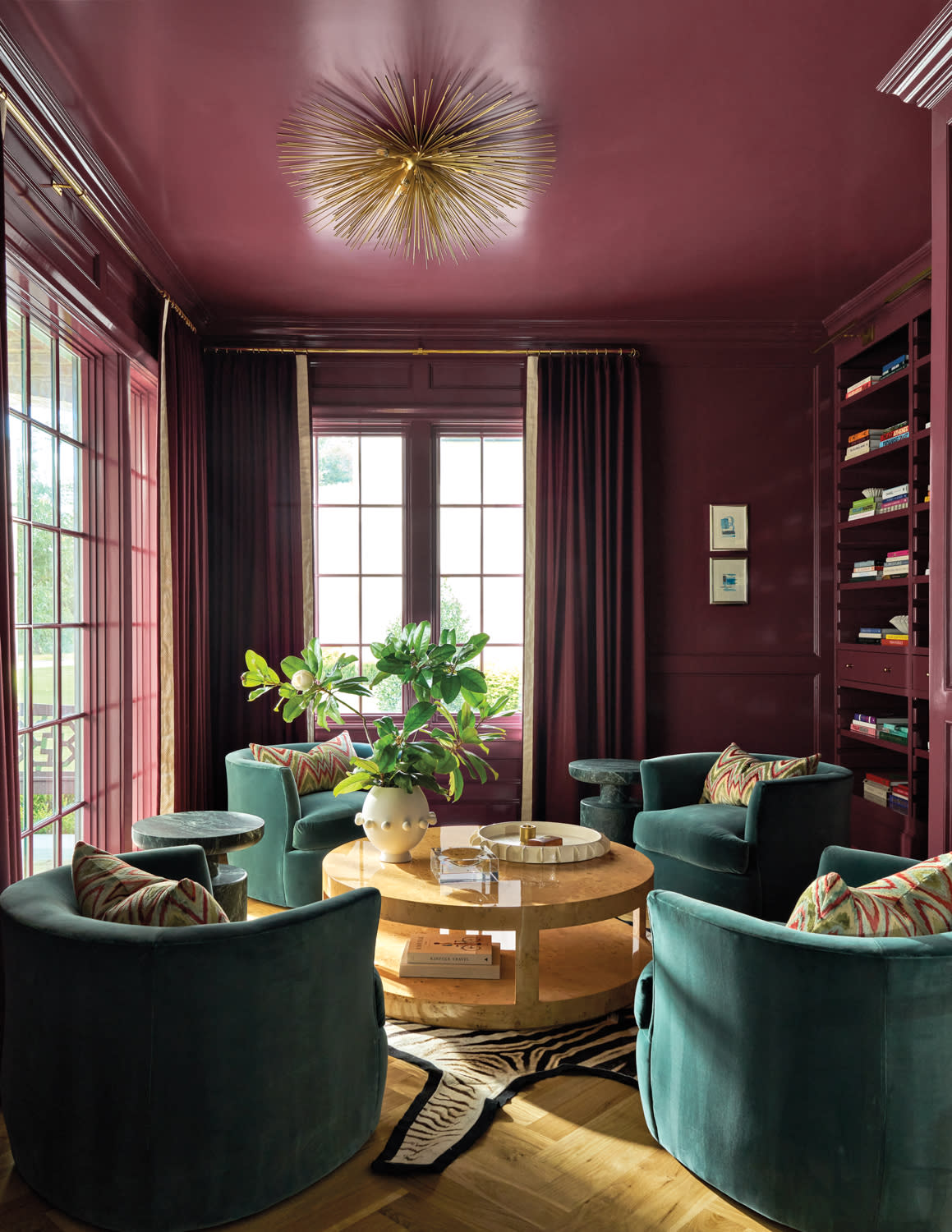 Elegant room with deep burgundy walls, teal velvet chairs, and a round wooden table. A large plant sits on the table beneath a gold spiky chandelier.