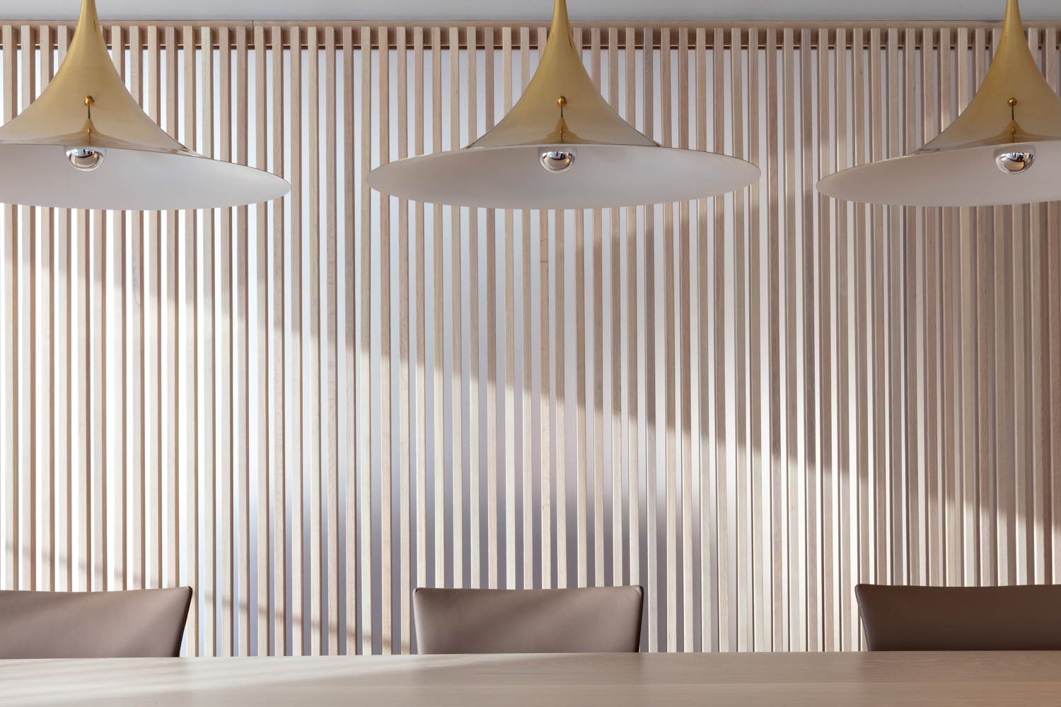 Wood-slated wall behind dining table with three hat-shaped pendants hanging above by Rodrigo Albir