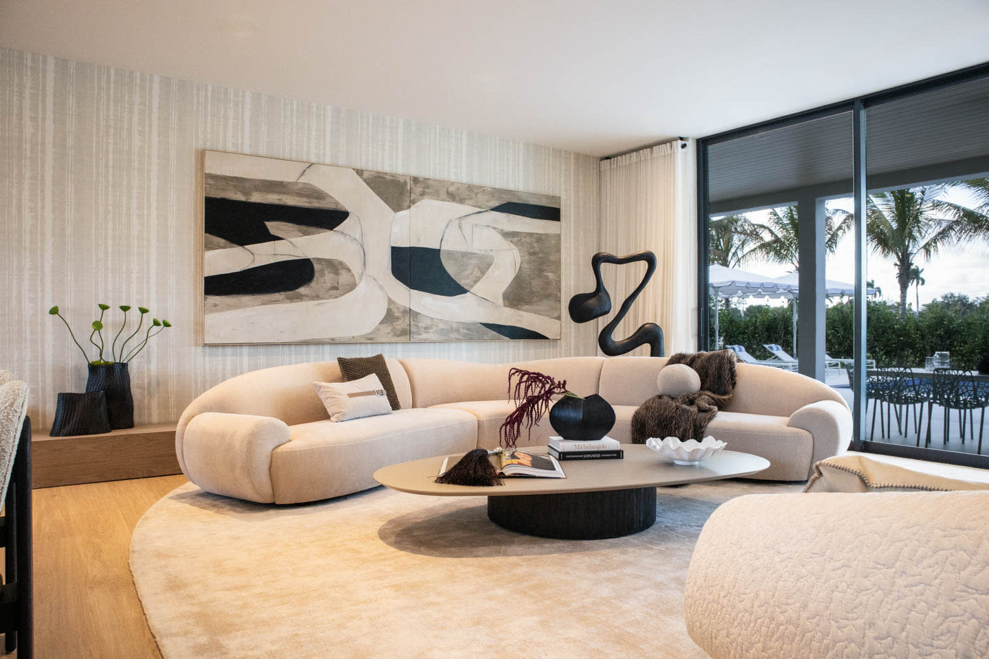 Modern living room with a beige, curved sofa, abstract artwork, and a circular coffee table. Large window reveals palm trees, creating a serene vibe.