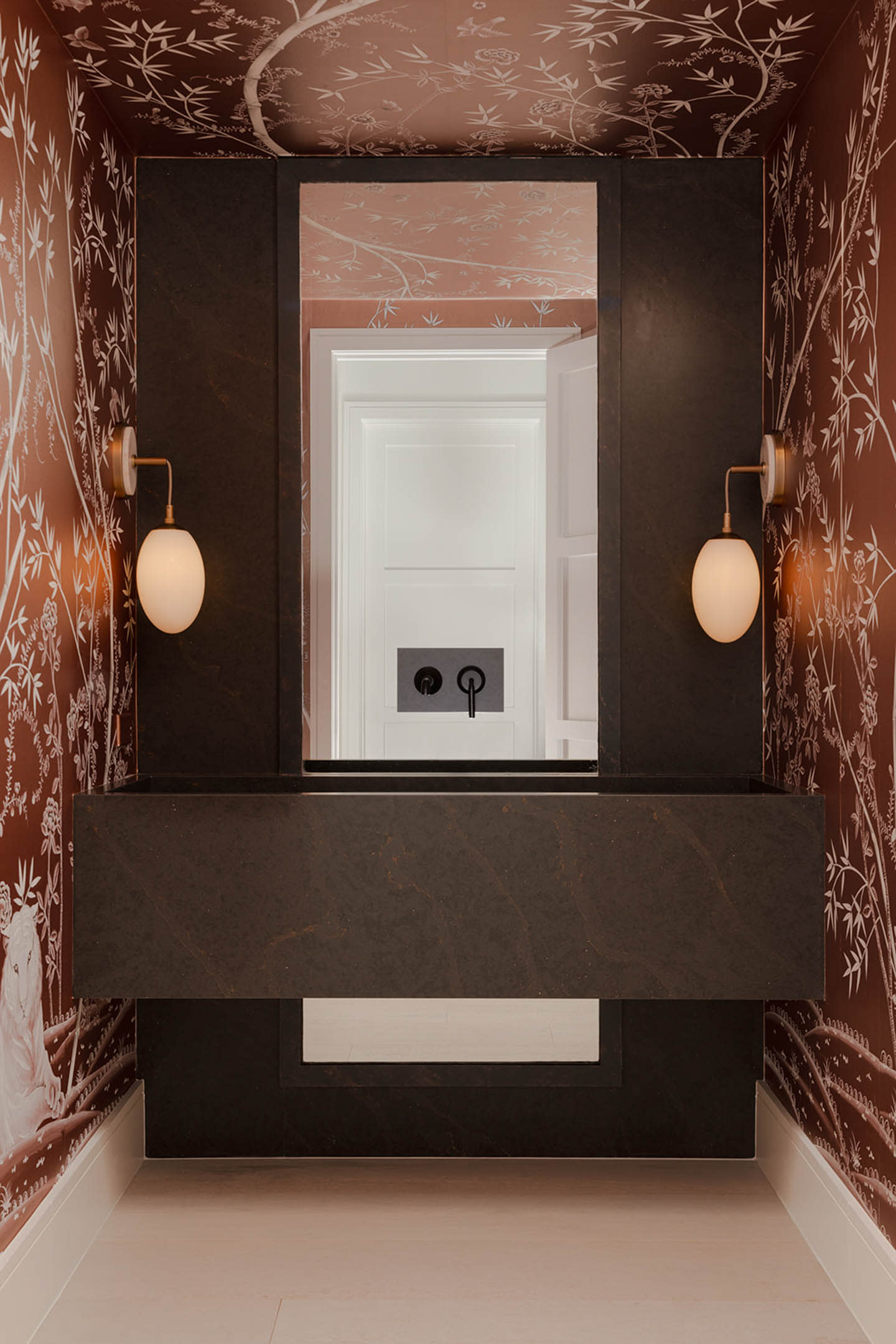 A small, elegant powder room with dark stone vanity and mirror. Wall sconces provide soft lighting. Brown wallpaper features white floral patterns.