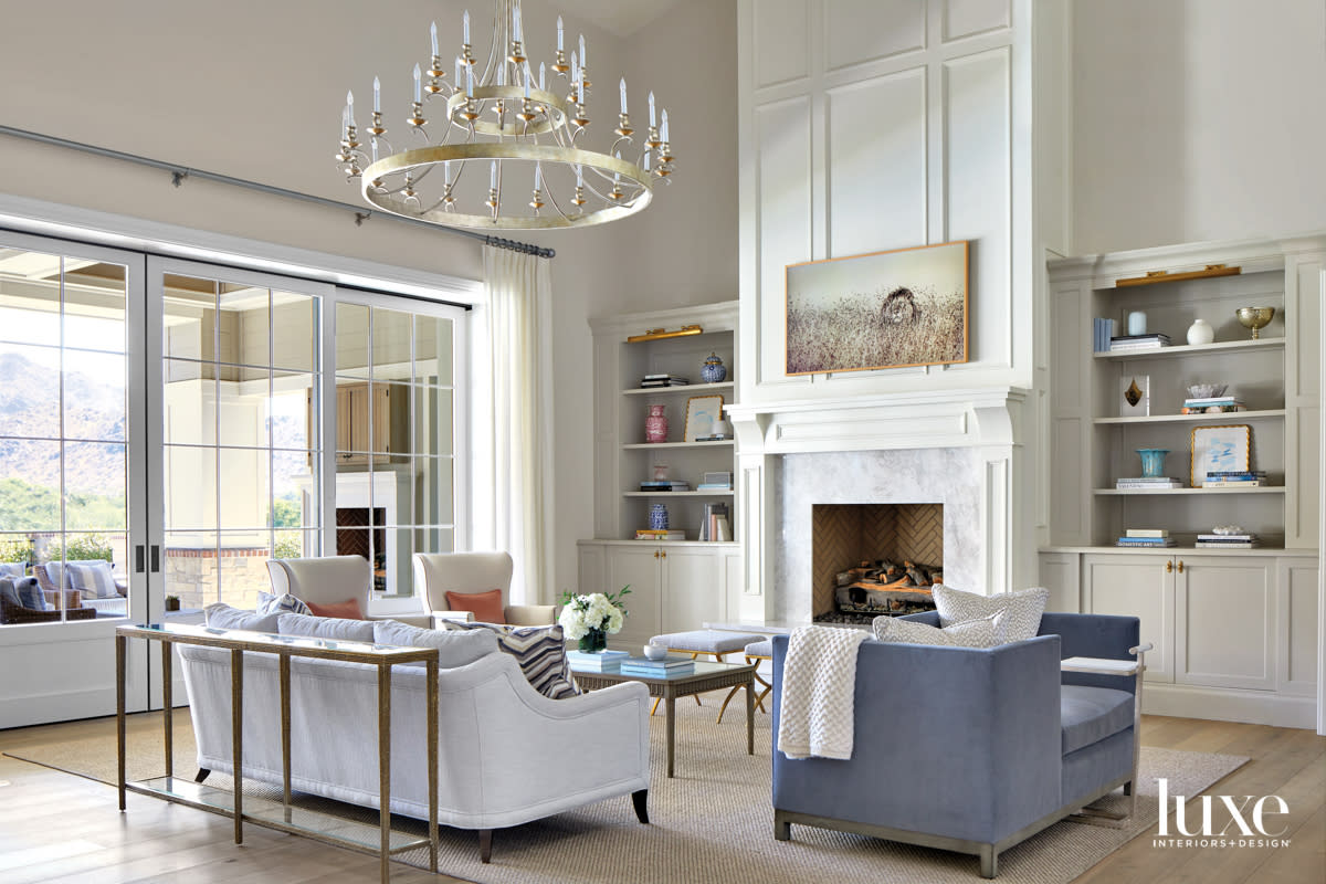 A white living room with a blue tete-a-tete, white couch, fireplace and chandelier.